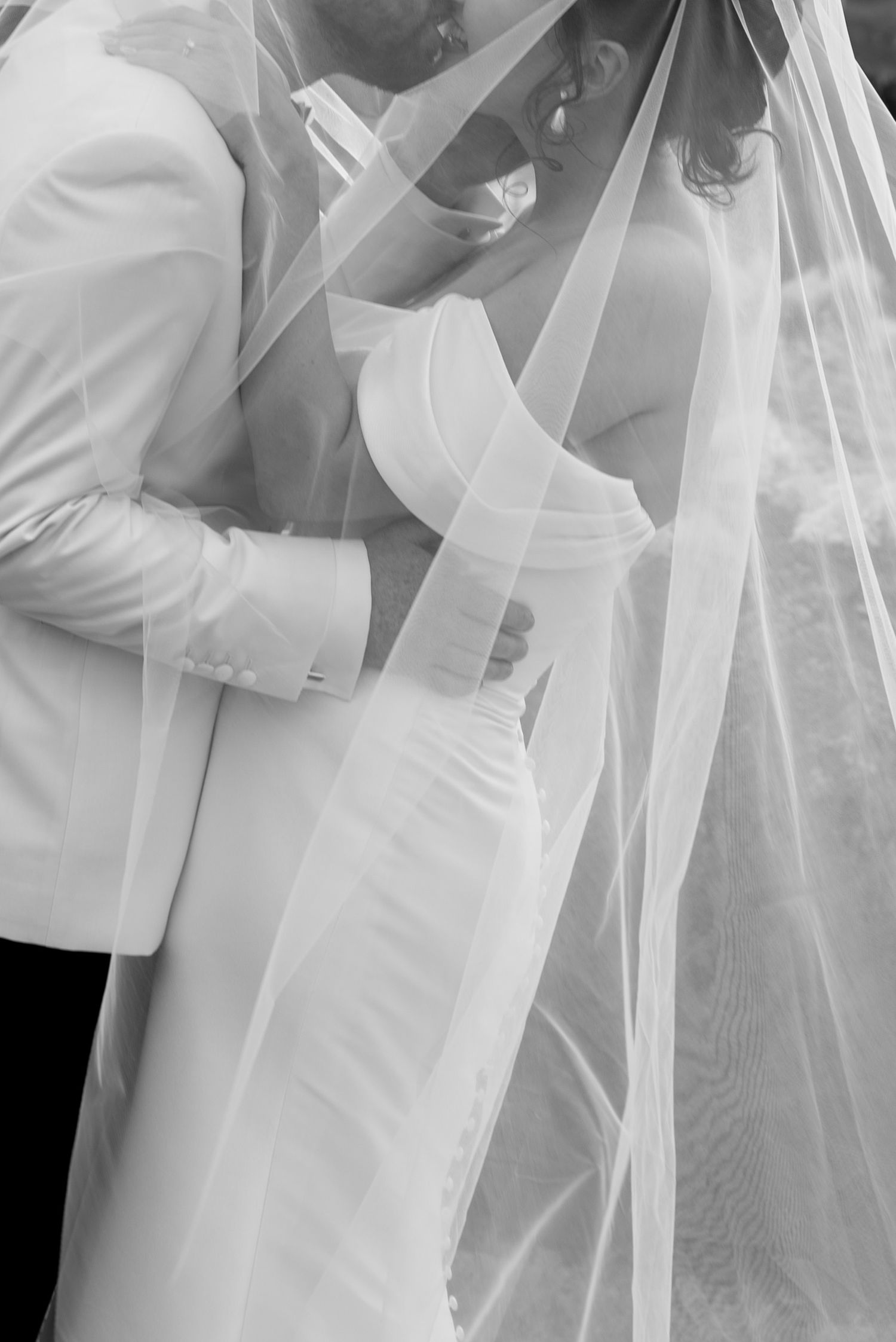 Black and white photo of a bride and groom sharing an intimate embrace under a flowing wedding veil.