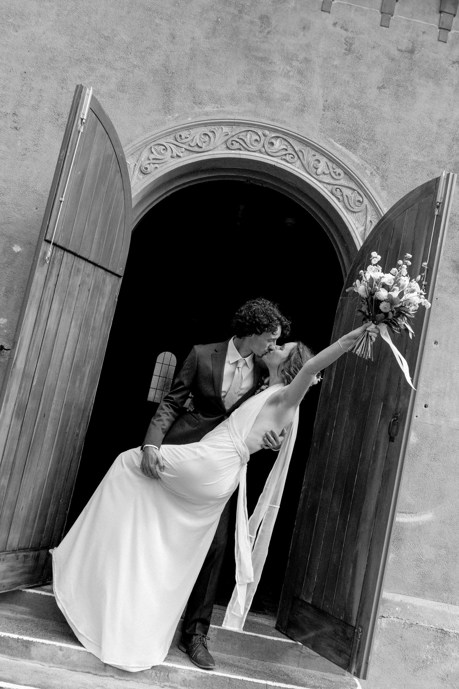 Black and white photo of a bride and groom sharing a romantic dip kiss in an ornate church doorway, bouquet in hand.