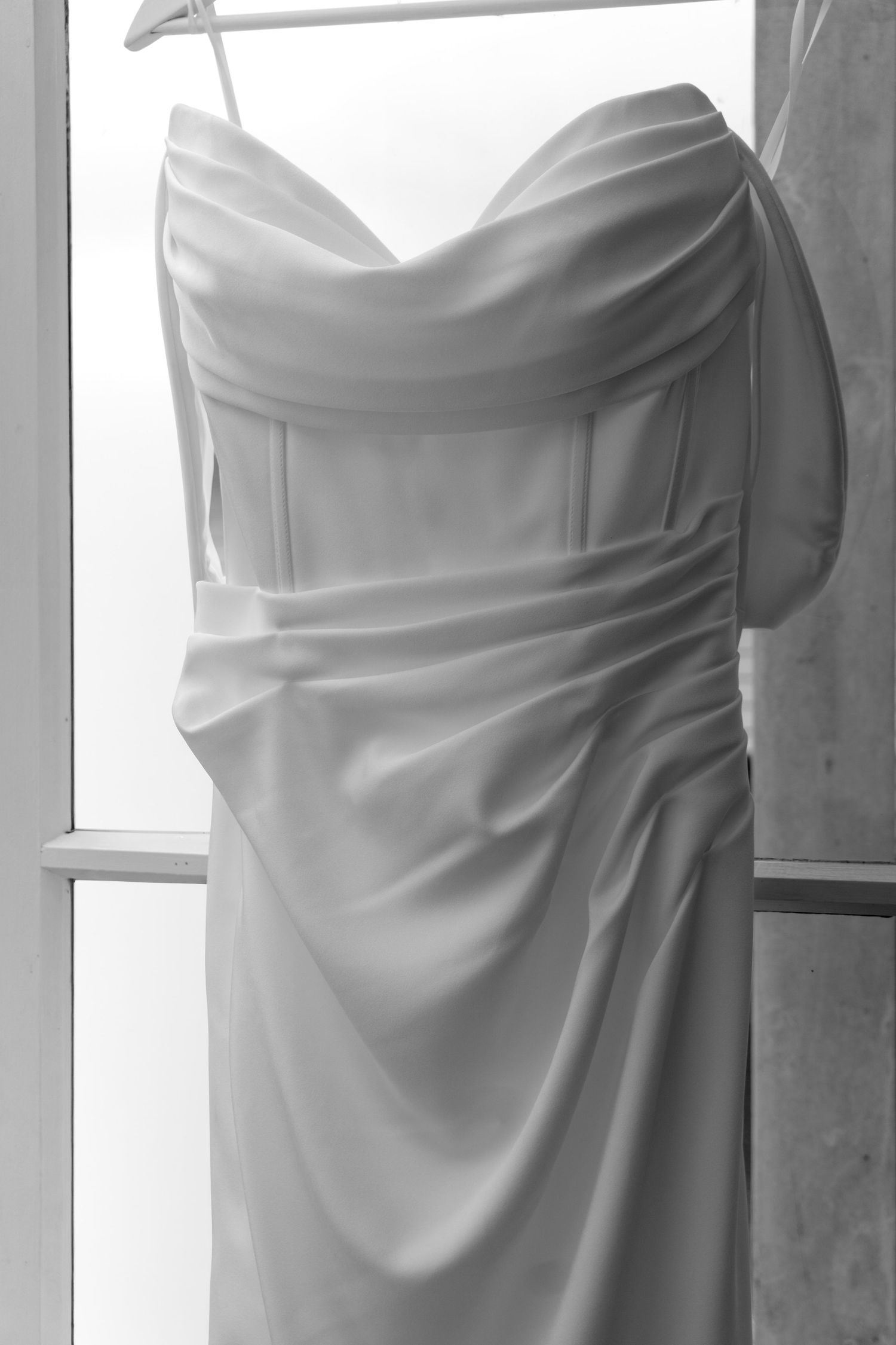 Black and white photo of an elegant strapless wedding dress with ruched satin bodice hanging by a window.