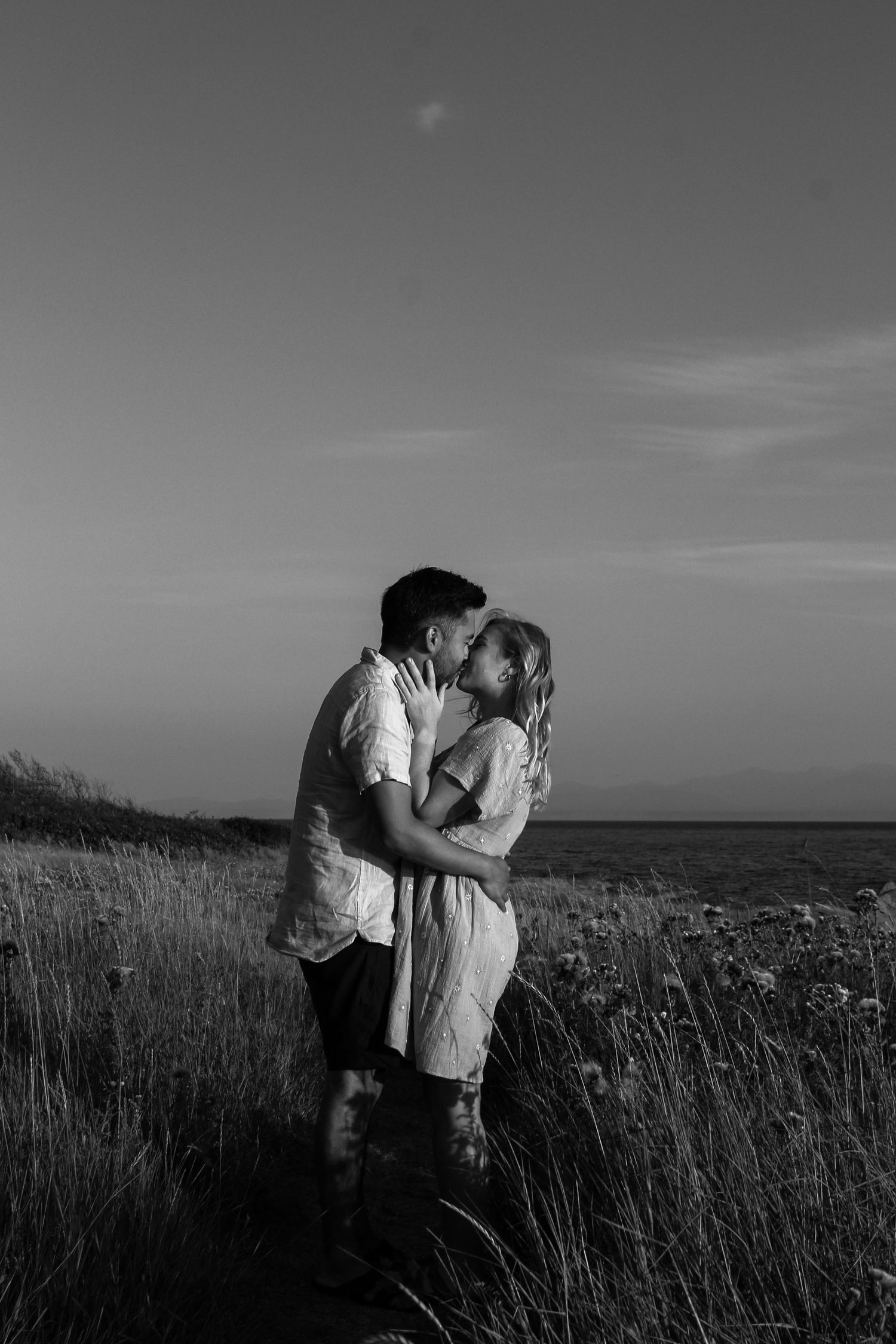 Black and white photo of couple embracing in tall grass with ocean horizon.