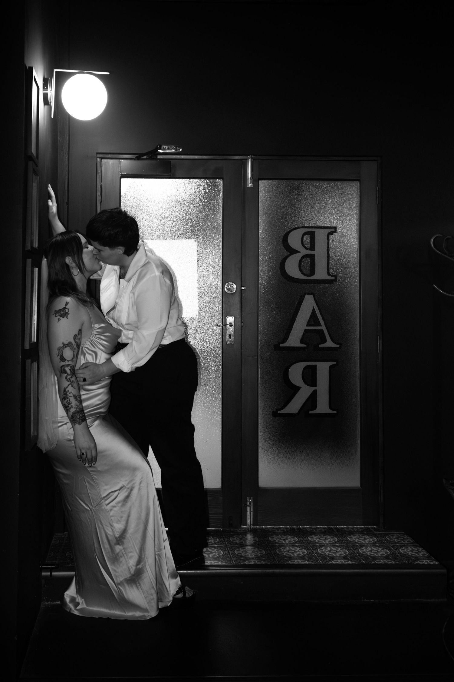 Black and white photo of a couple sharing a romantic kiss outside a bar entrance at night.