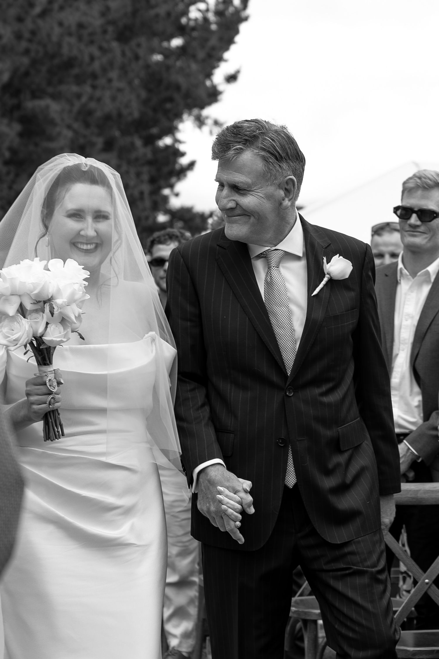 Black and white photo of a smiling bride in veil holding bouquet walking with groom in suit at outdoor wedding ceremony.