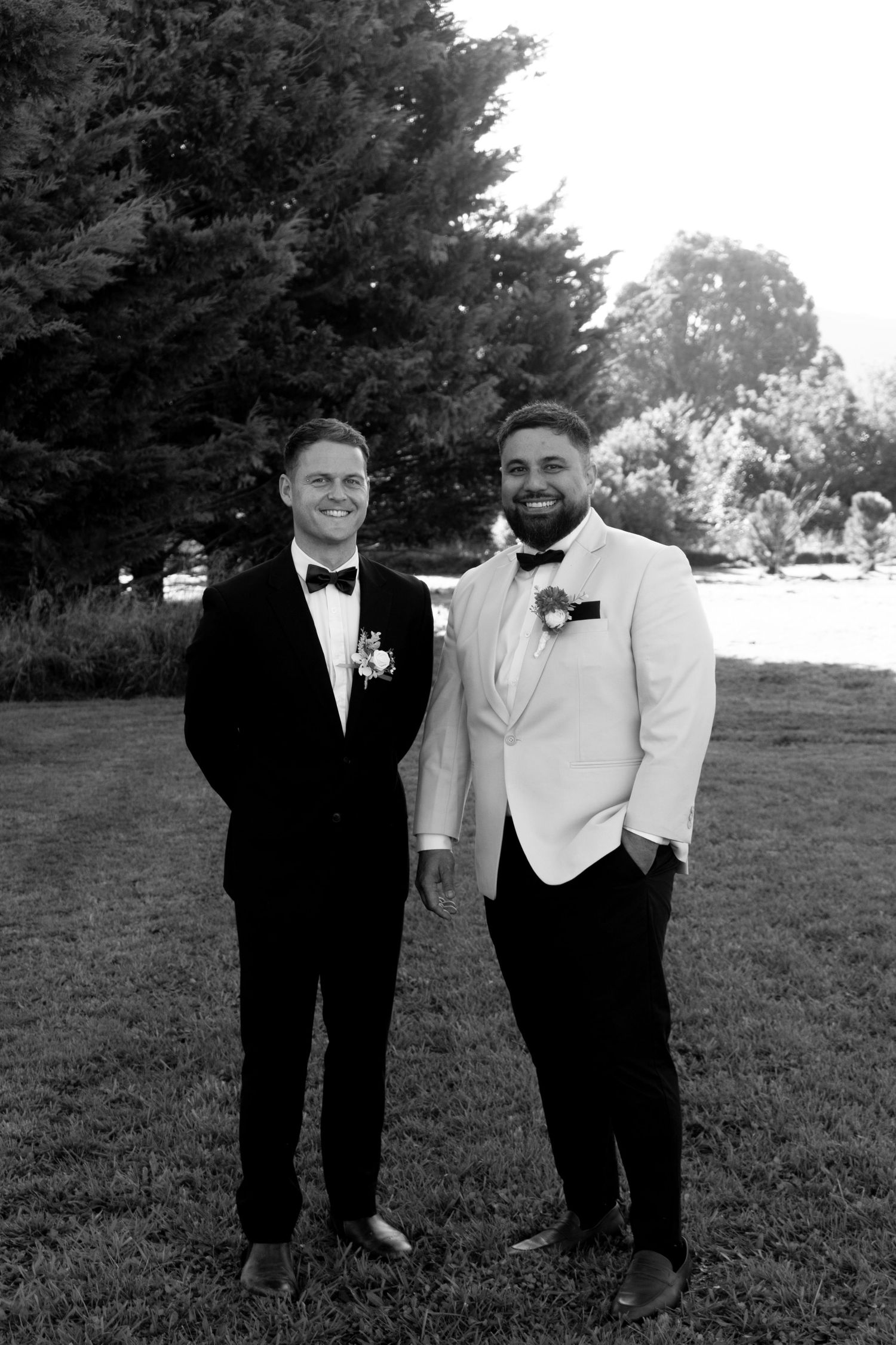 Two grooms in black and white tuxedos with boutonnieres pose outdoors on grass with trees in background.