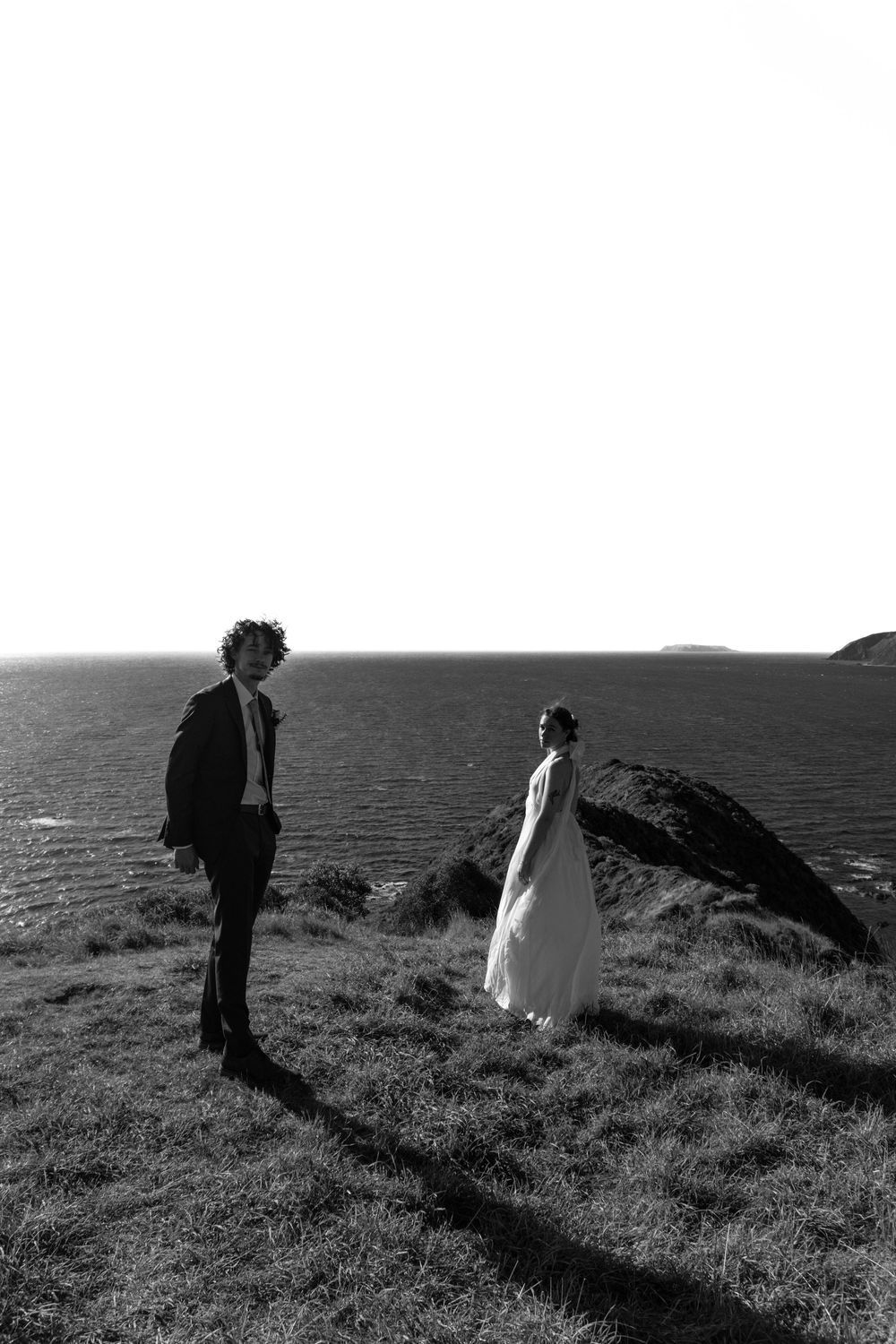 Black and white photo of a bride and groom standing on a coastal cliff overlooking the ocean on their wedding day.