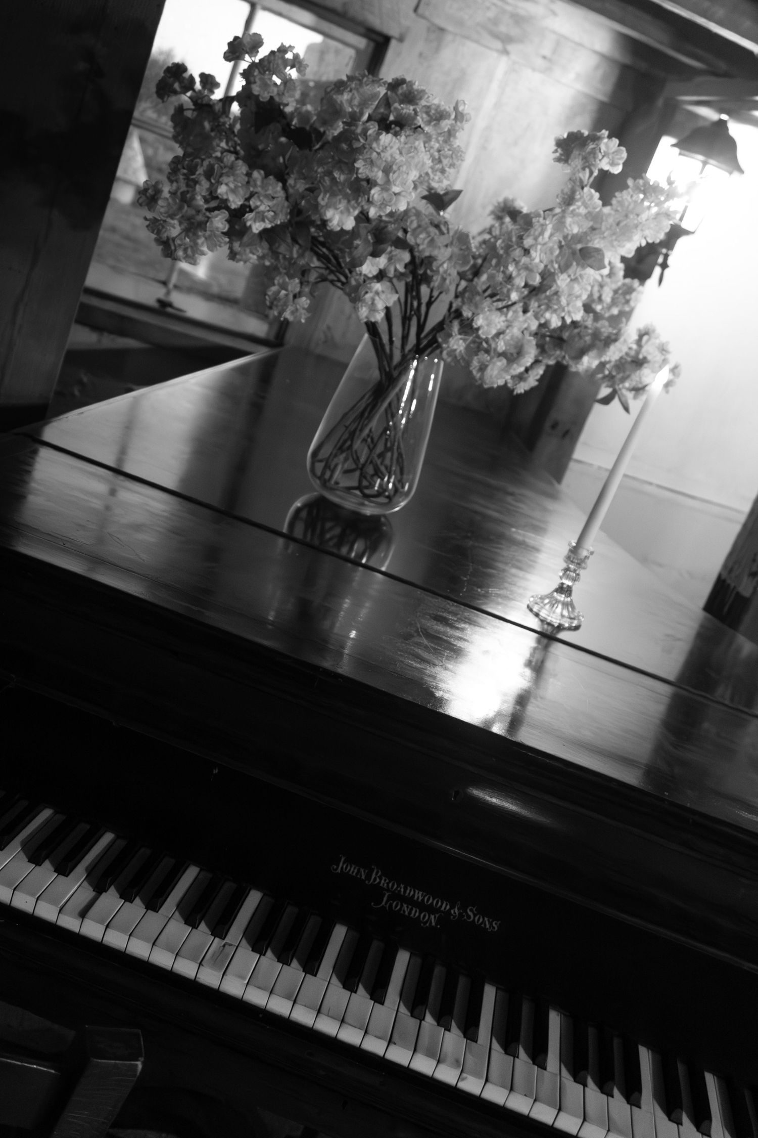 Black and white photo of a grand piano with floral arrangement in glass vase and candle on its polished surface.