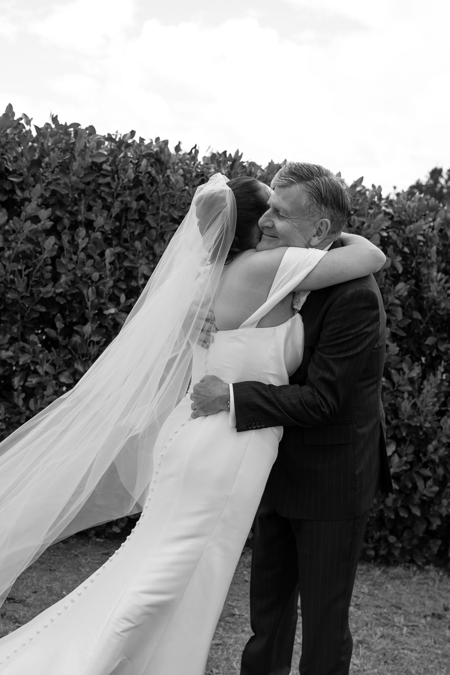 Black and white photo of a bride in a flowing veil embracing a man in a suit near lush greenery.