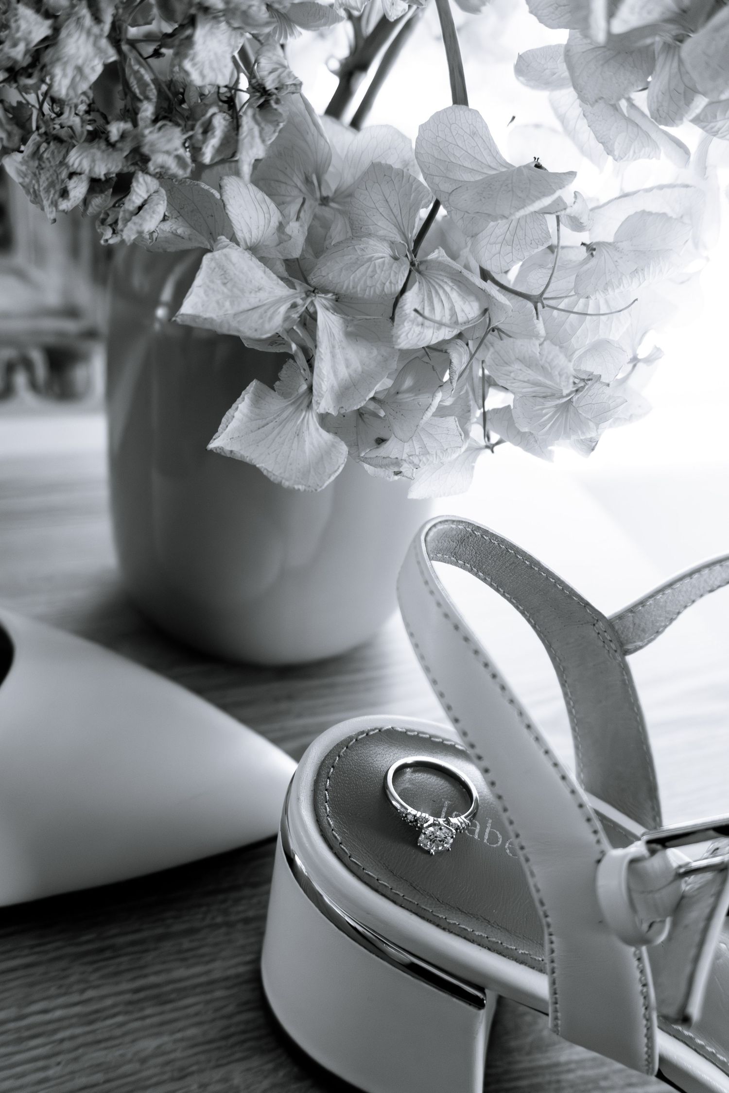 Black and white photo of a diamond engagement ring resting on a white bridal heel beside a vase of hydrangeas.