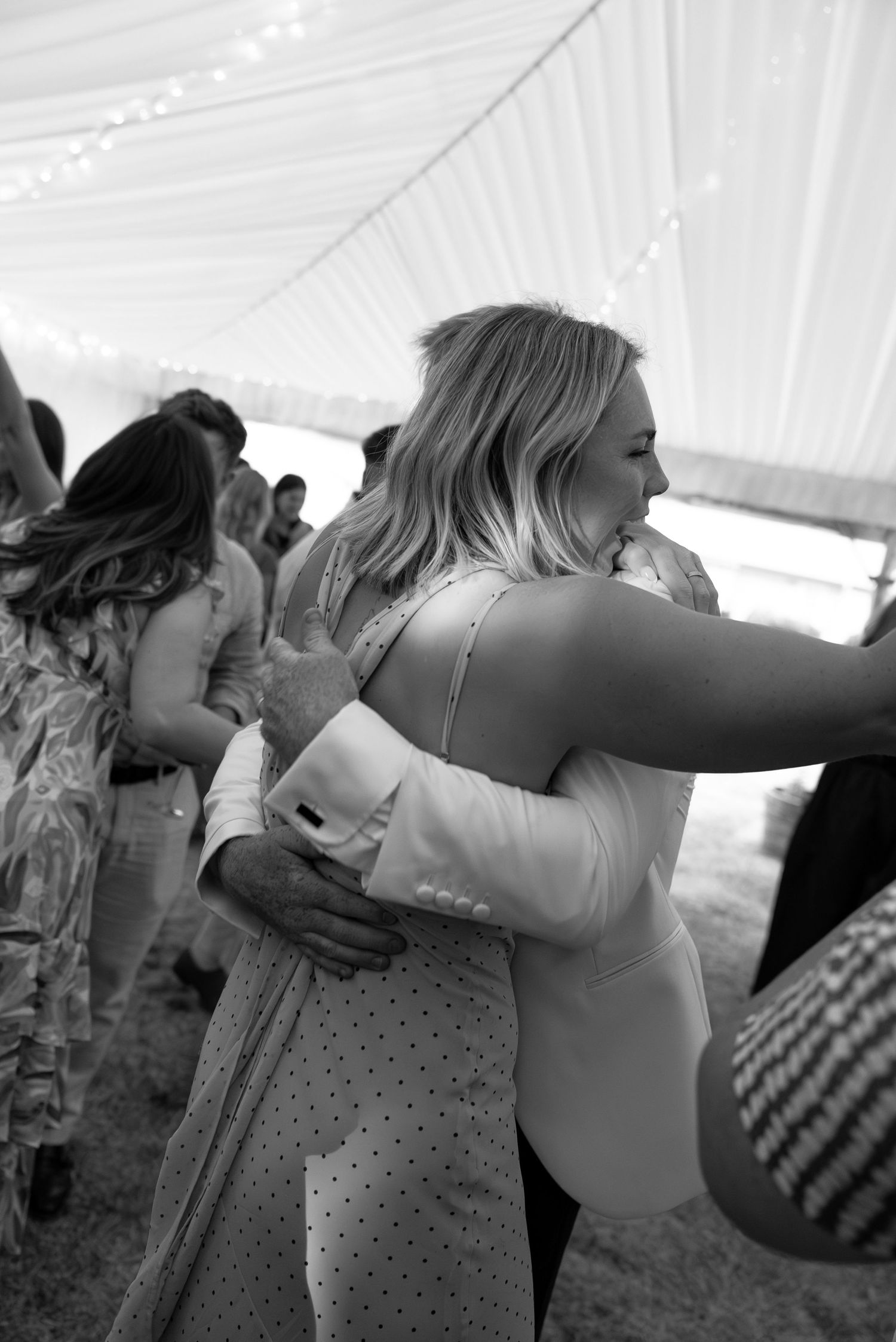 Black and white photo of guests hugging and dancing under a tent at a wedding reception.
