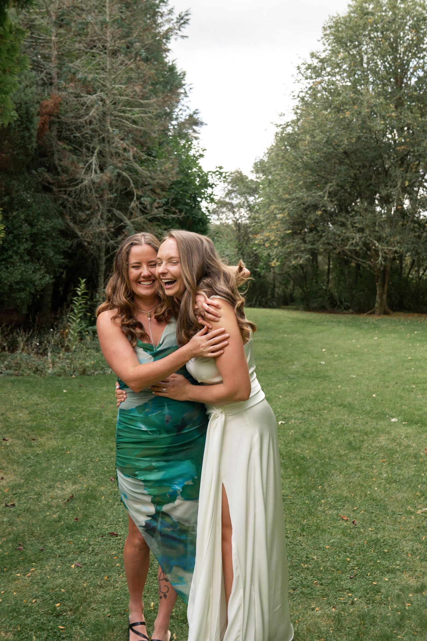 Two women in elegant dresses laughing and hugging outdoors on green grass with trees in the background.