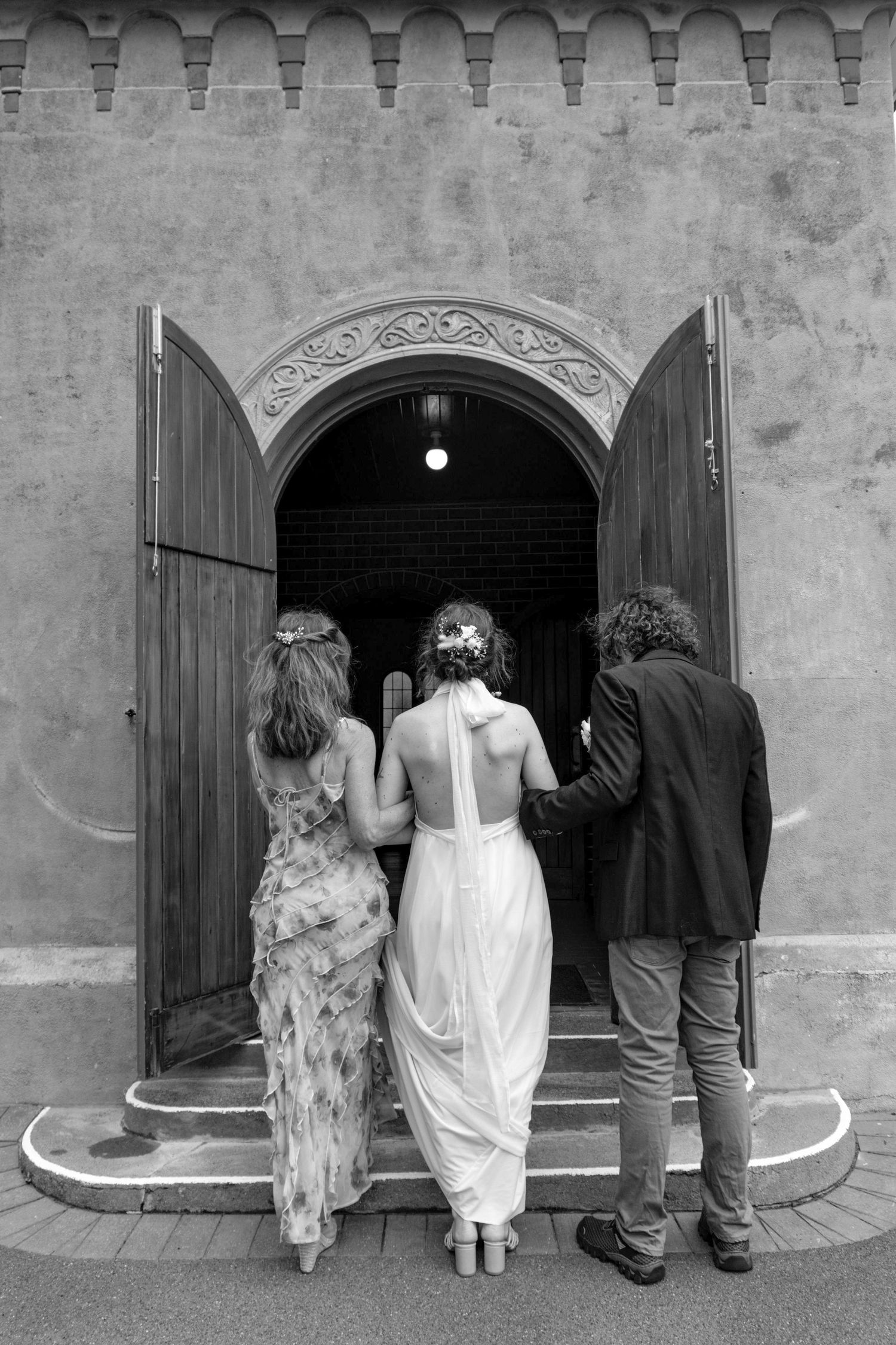 Black and white photo of a bride in white gown entering a church with two guests through an ornate arched doorway.
