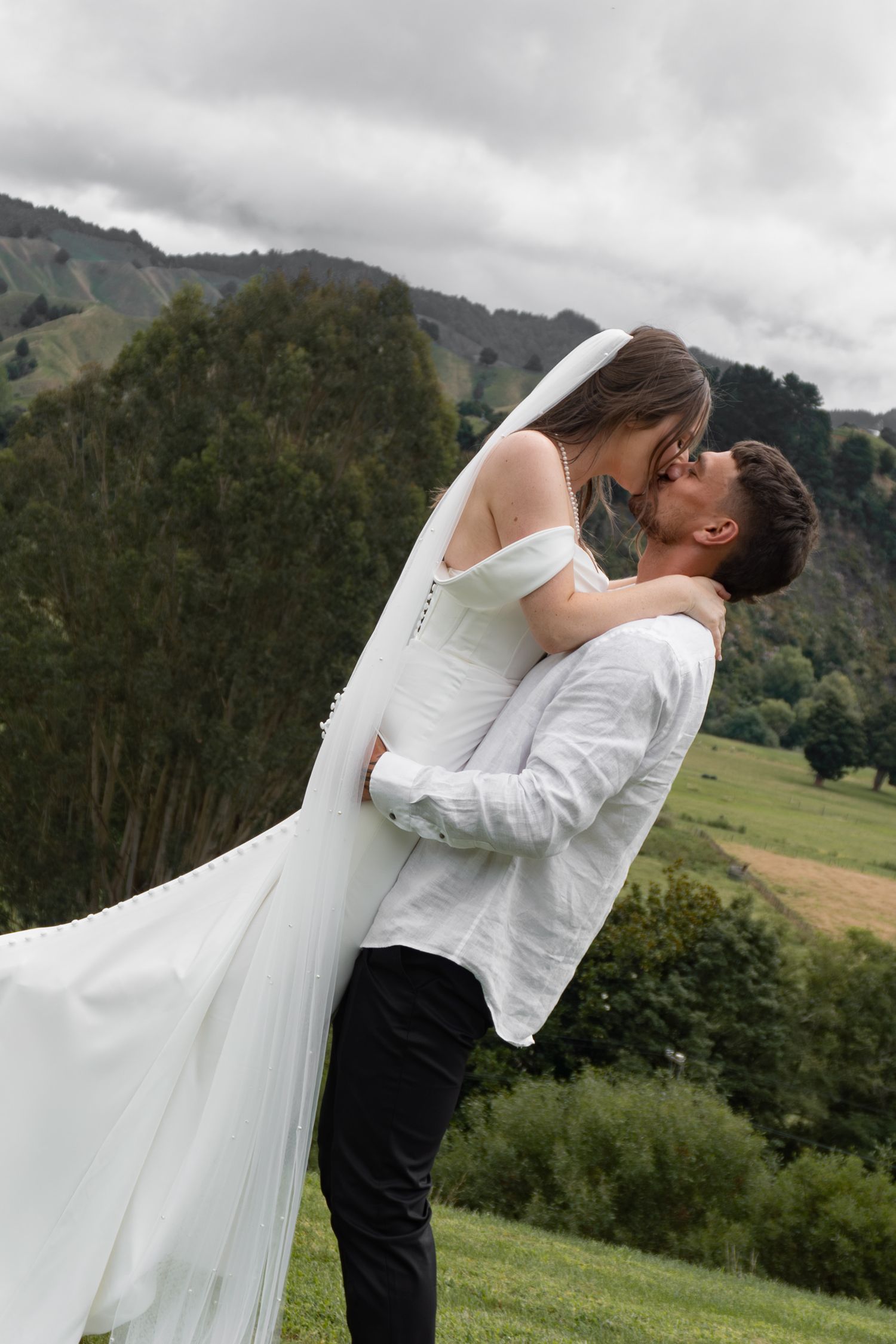 Groom lifting bride in white dress and veil, sharing a kiss with mountain landscape backdrop.