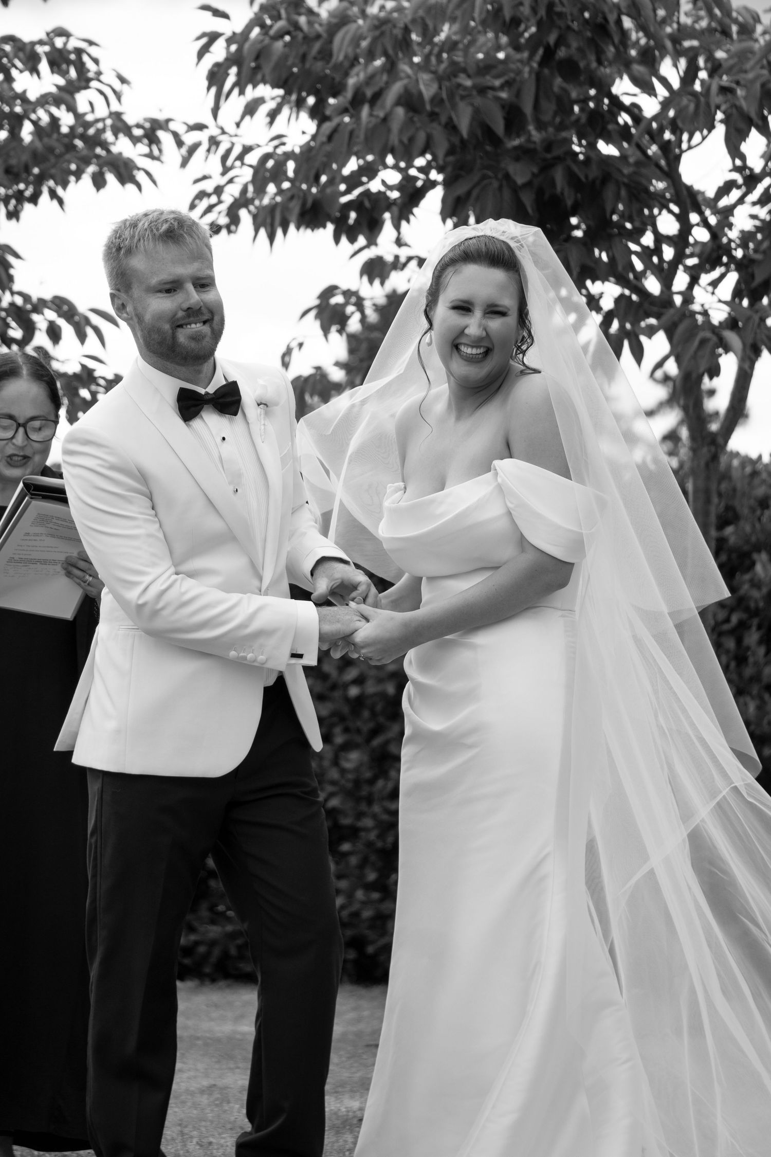 Black and white photo of a smiling bride and groom holding hands during an outdoor wedding ceremony.