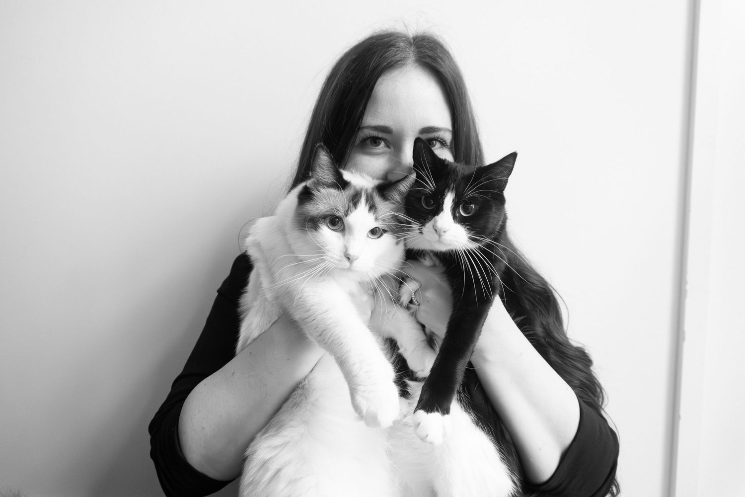 Black and white photo of a young woman holding two cats, one white and one black, close to her face.