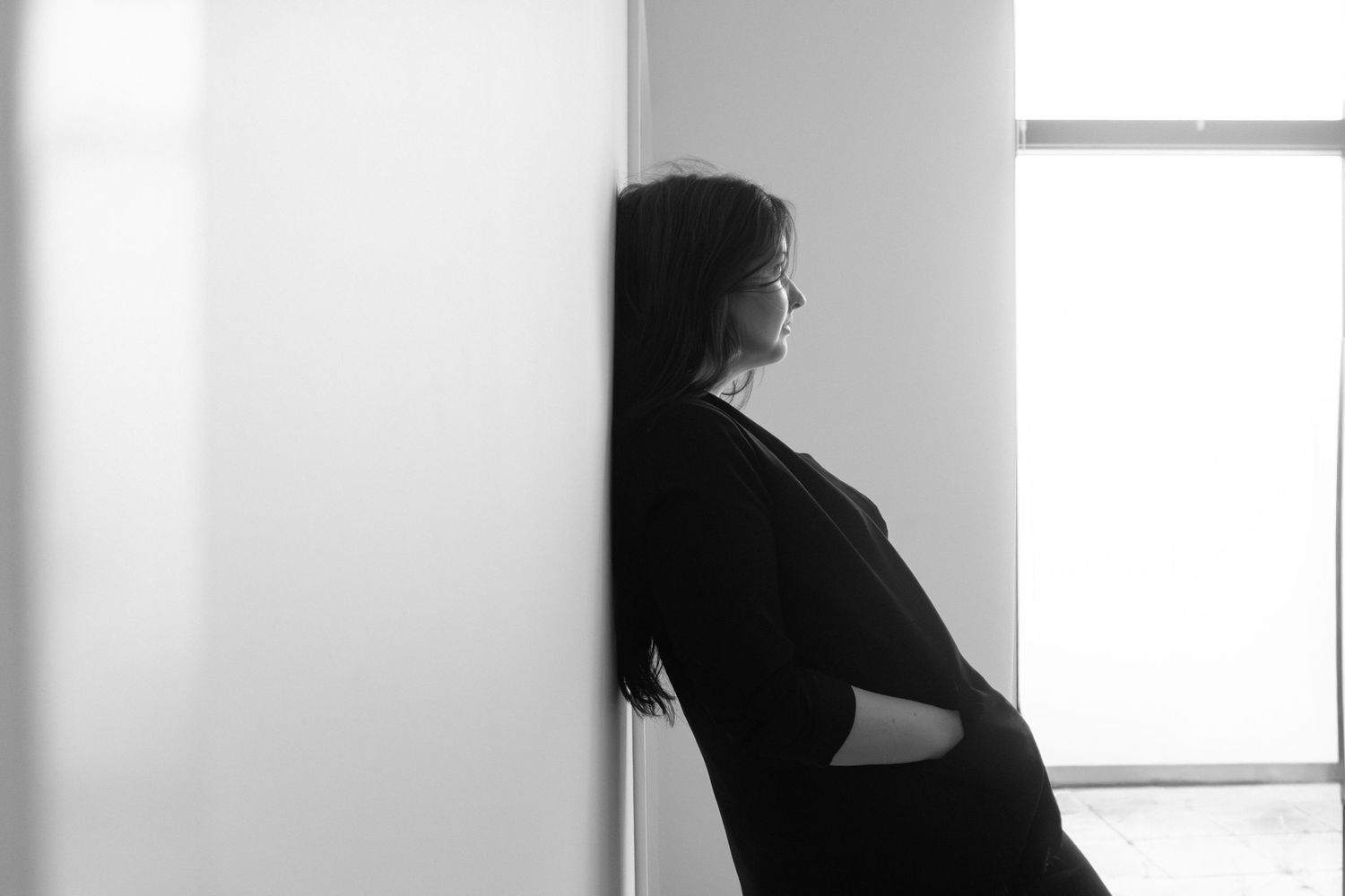 Black and white photo of a pregnant woman leaning against a wall, gazing toward a bright window.
