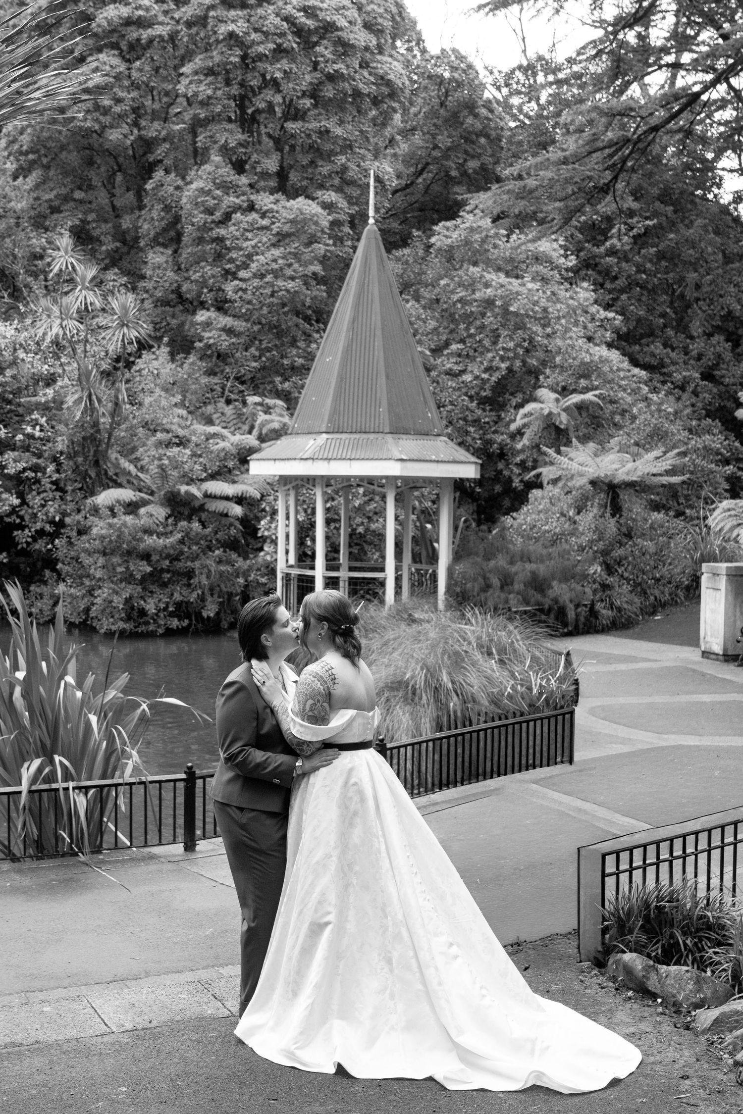 Black and white photo of a bride and groom kissing in a garden with a gazebo and lush trees in the background.