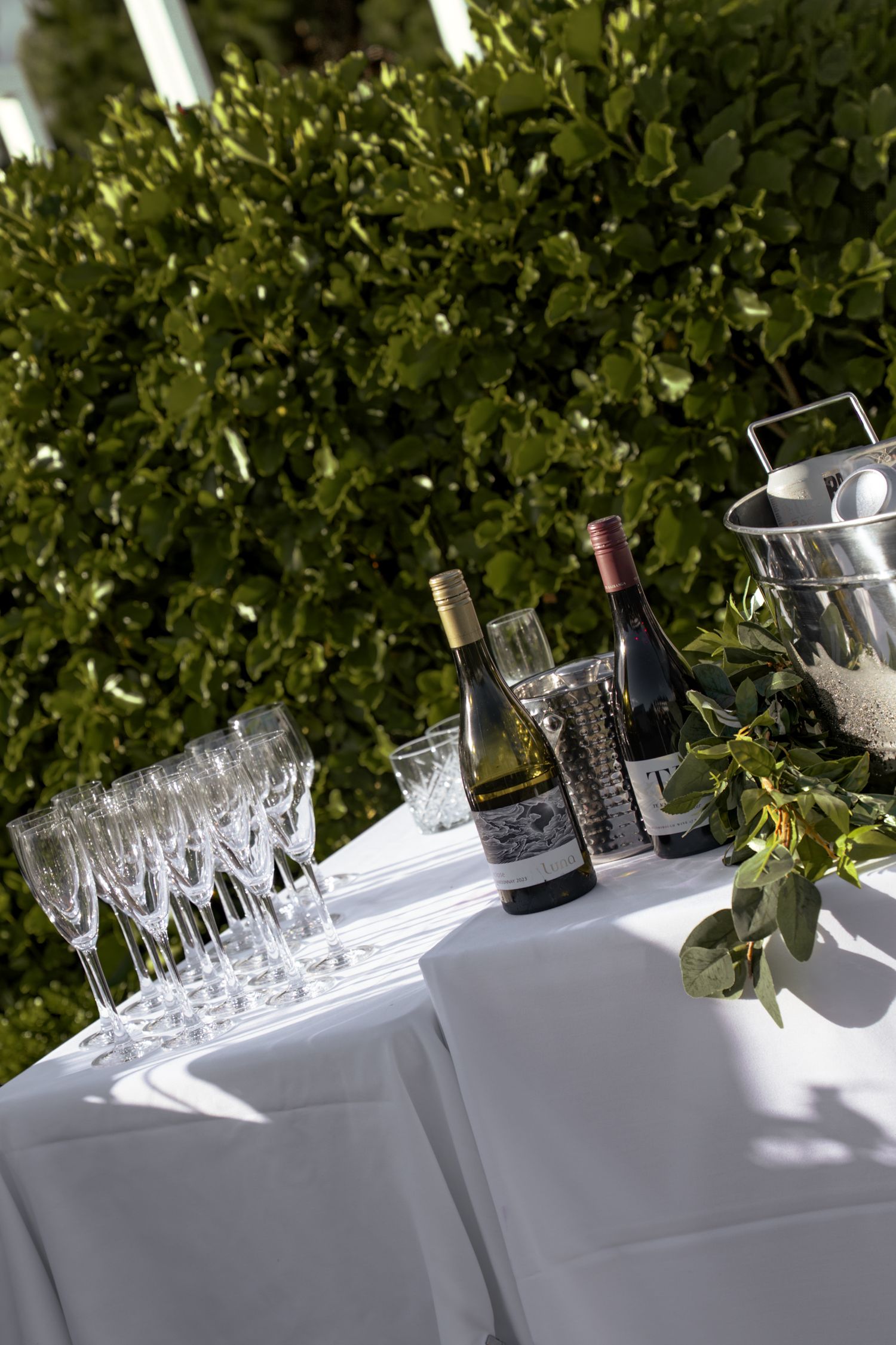Elegant outdoor bar setup with crystal wine glasses, two wine bottles, and a silver ice bucket on a white linen table.