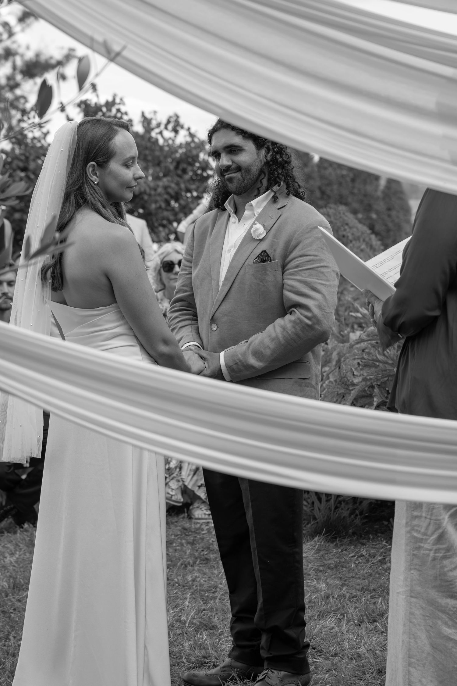 Black and white photo of a bride and groom holding hands at an outdoor wedding ceremony framed by flowing white drapes.