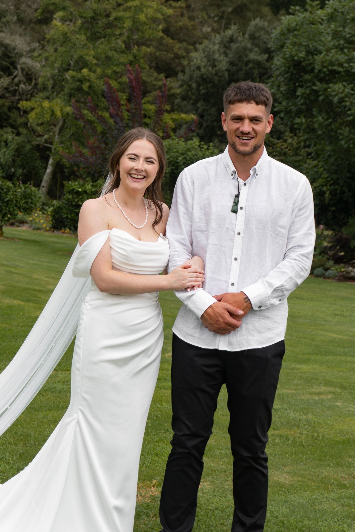 Bride in white gown and groom in white shirt pose together smiling on green lawn at outdoor wedding.