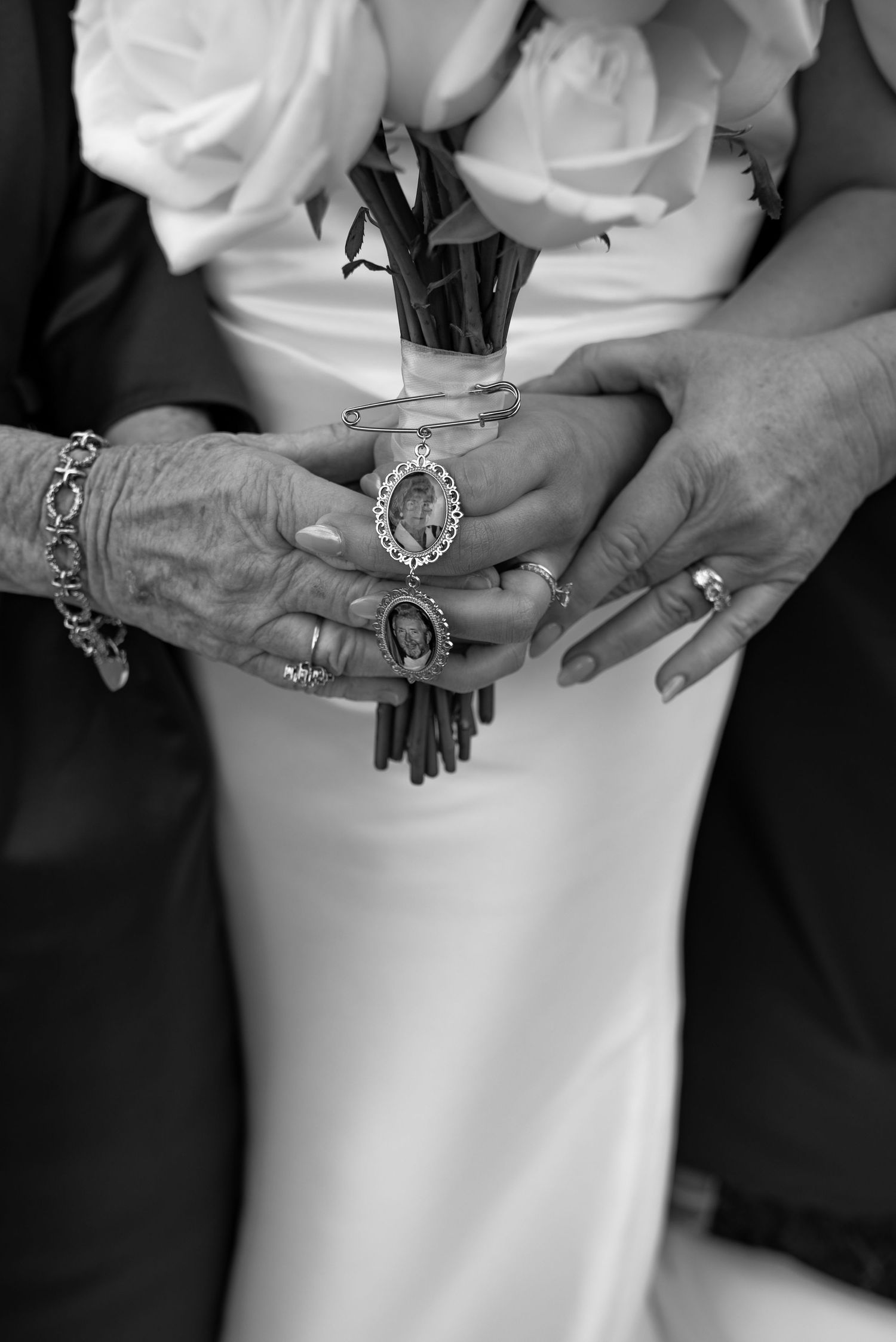 Black and white wedding photo of hands holding a rose bouquet with memorial photo charms, featuring rings and a bracelet.