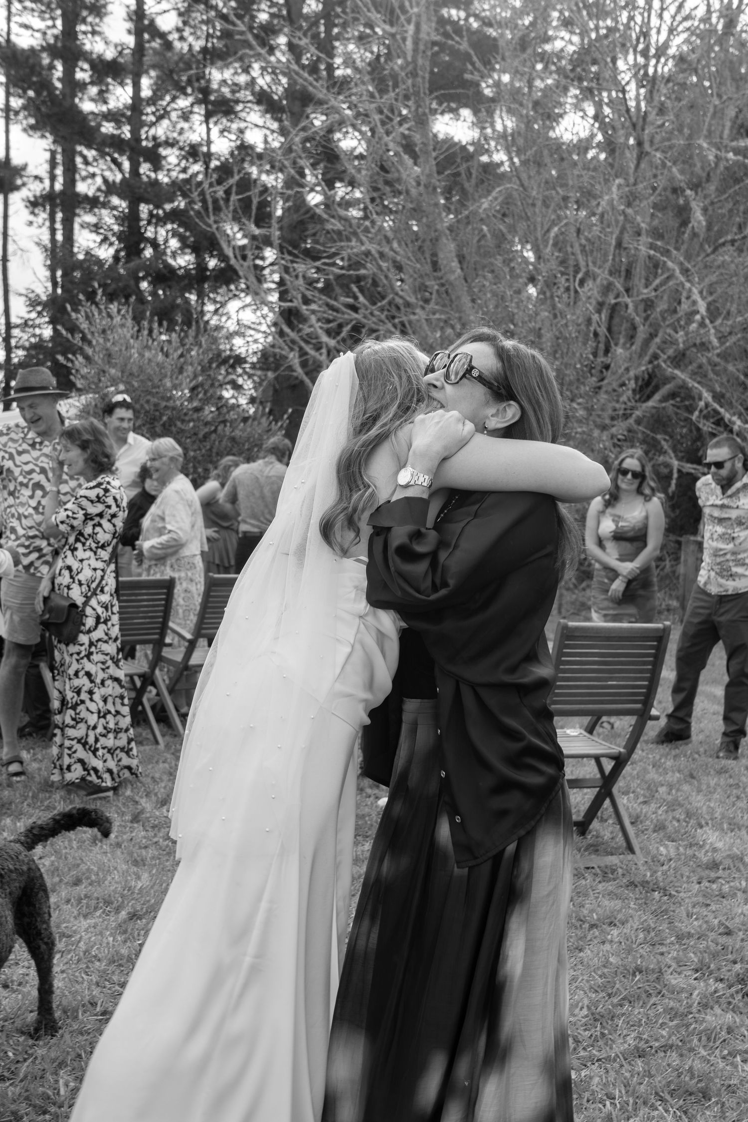 Black and white photo of a bride in a veil embracing a guest in a dark dress at an outdoor wedding ceremony.