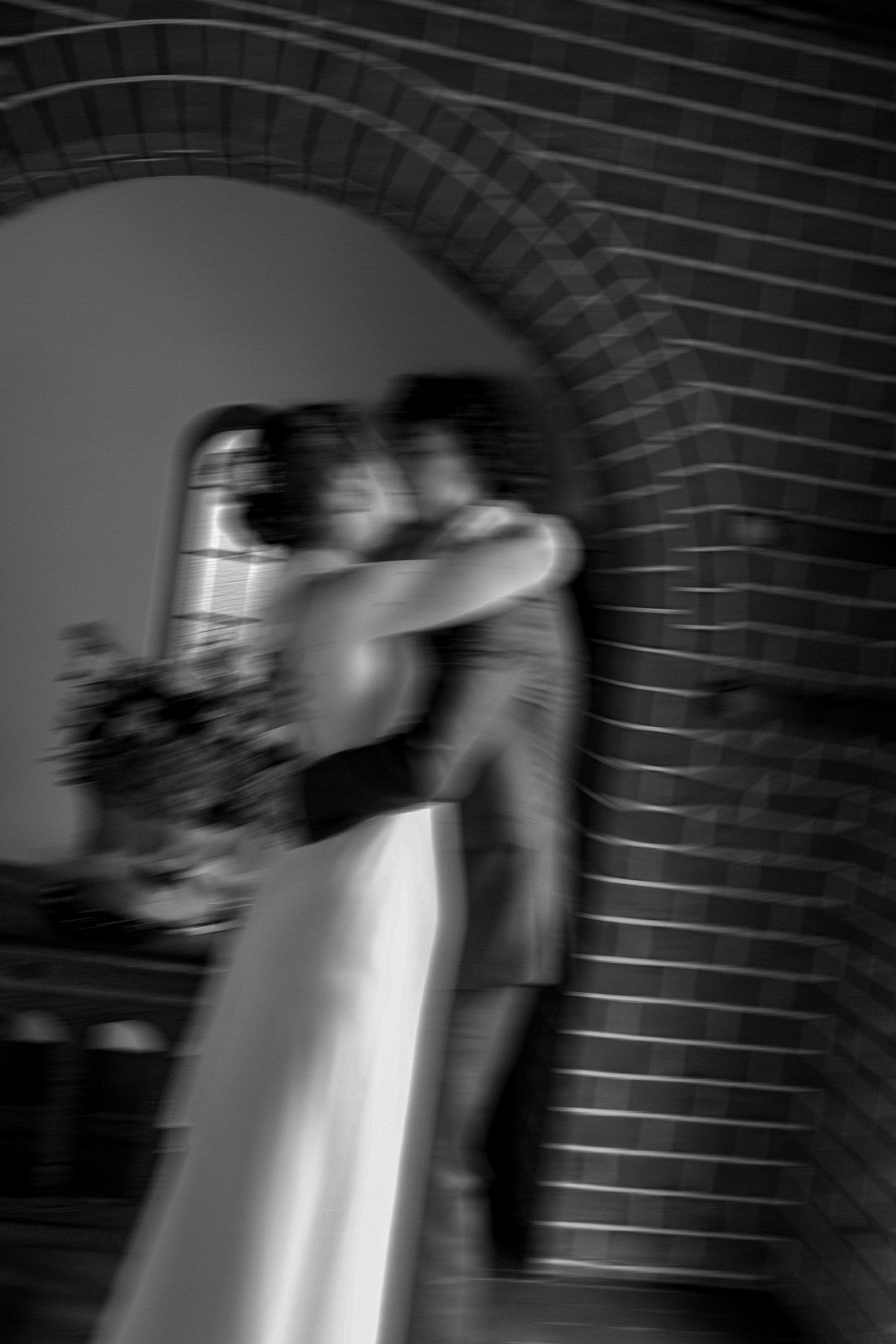 Black and white blurred photo of a bride and groom embracing under a brick arch at their wedding.