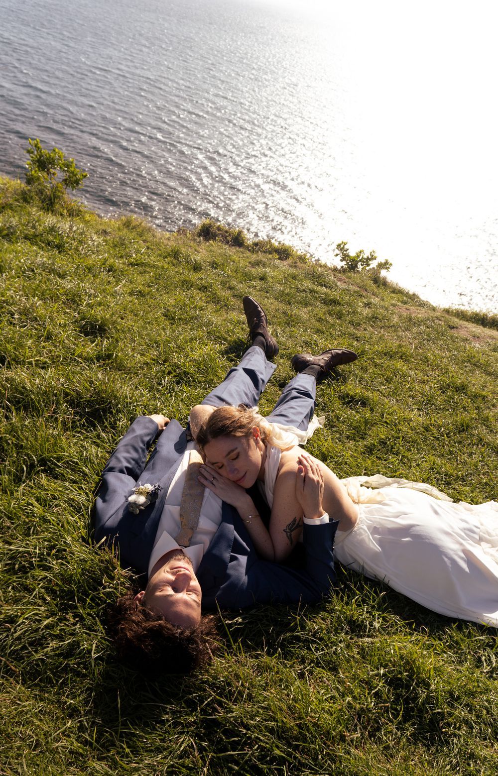 Bride and groom in wedding attire lying on a grassy hillside overlooking the ocean on their wedding day.