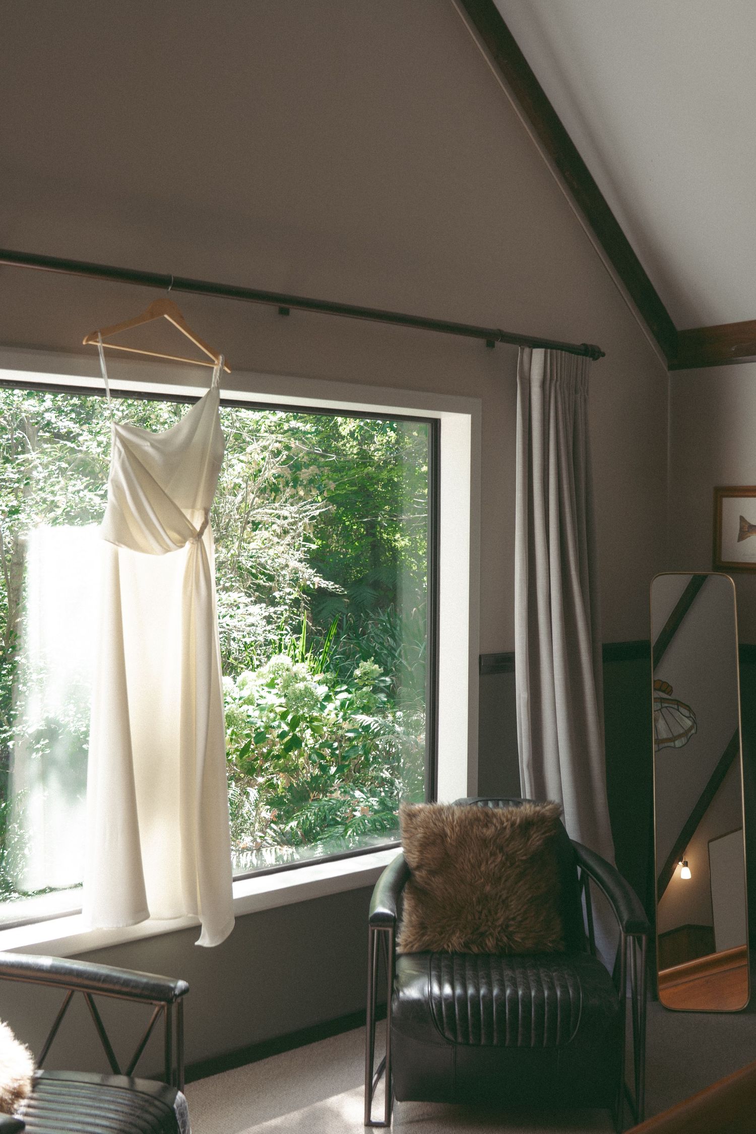 White wedding dress hanging on a wooden hanger in front of a large window with lush green trees outside.