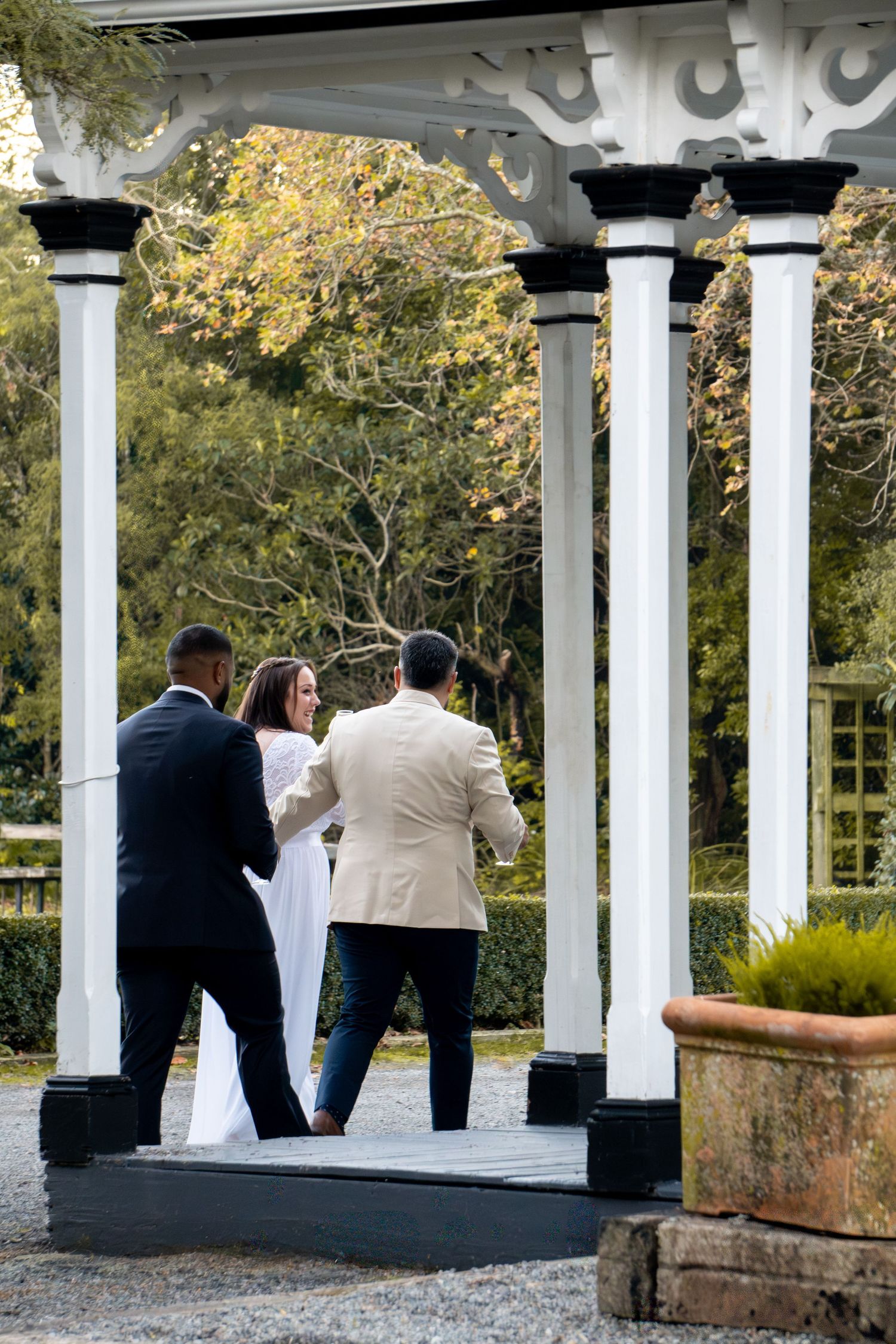 Three people stand under a white garden gazebo with columns, surrounded by lush greenery, during an outdoor ceremony.