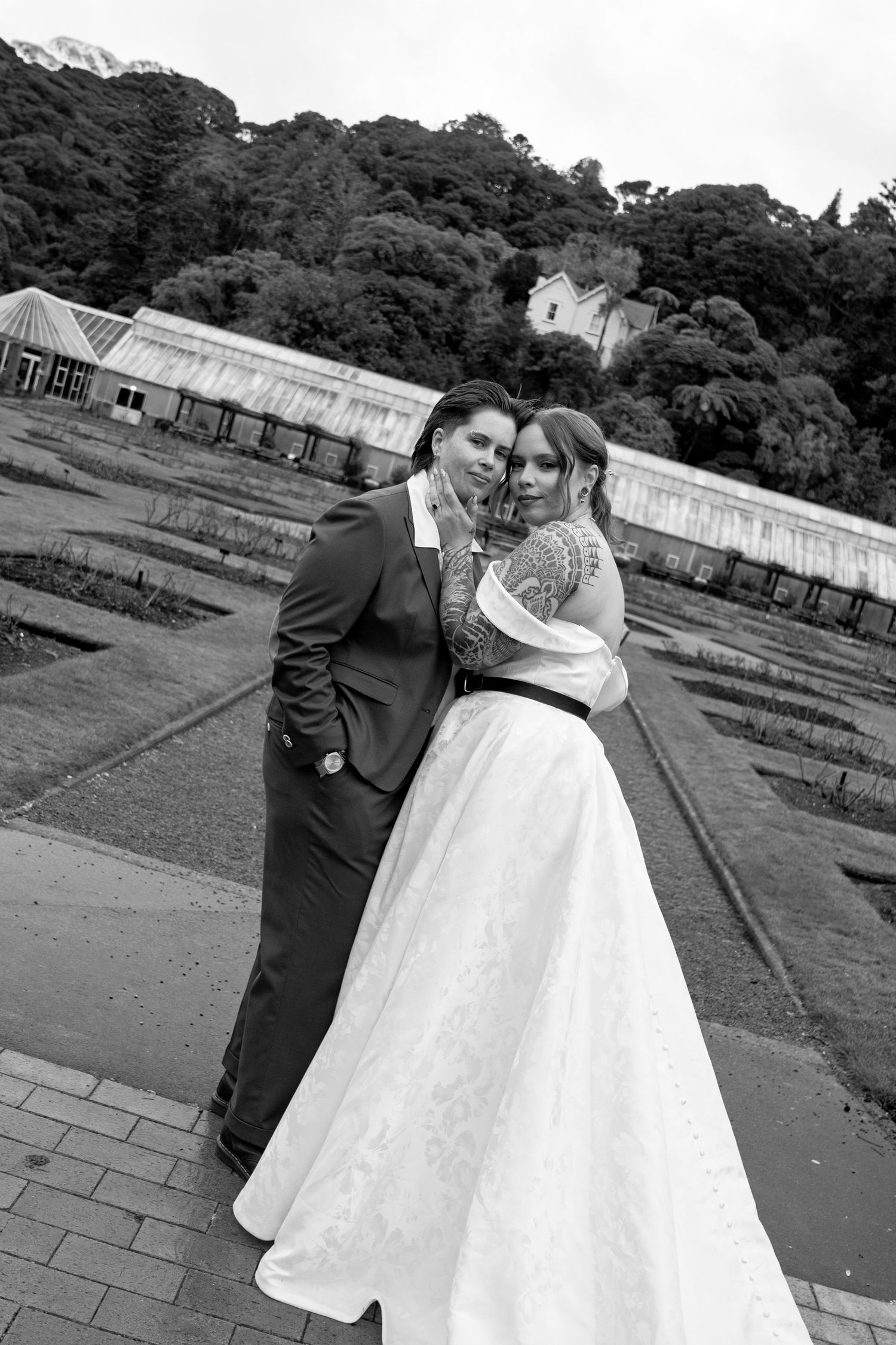 Black and white wedding photo of bride and groom embracing outdoors with lush trees and greenhouse in background.