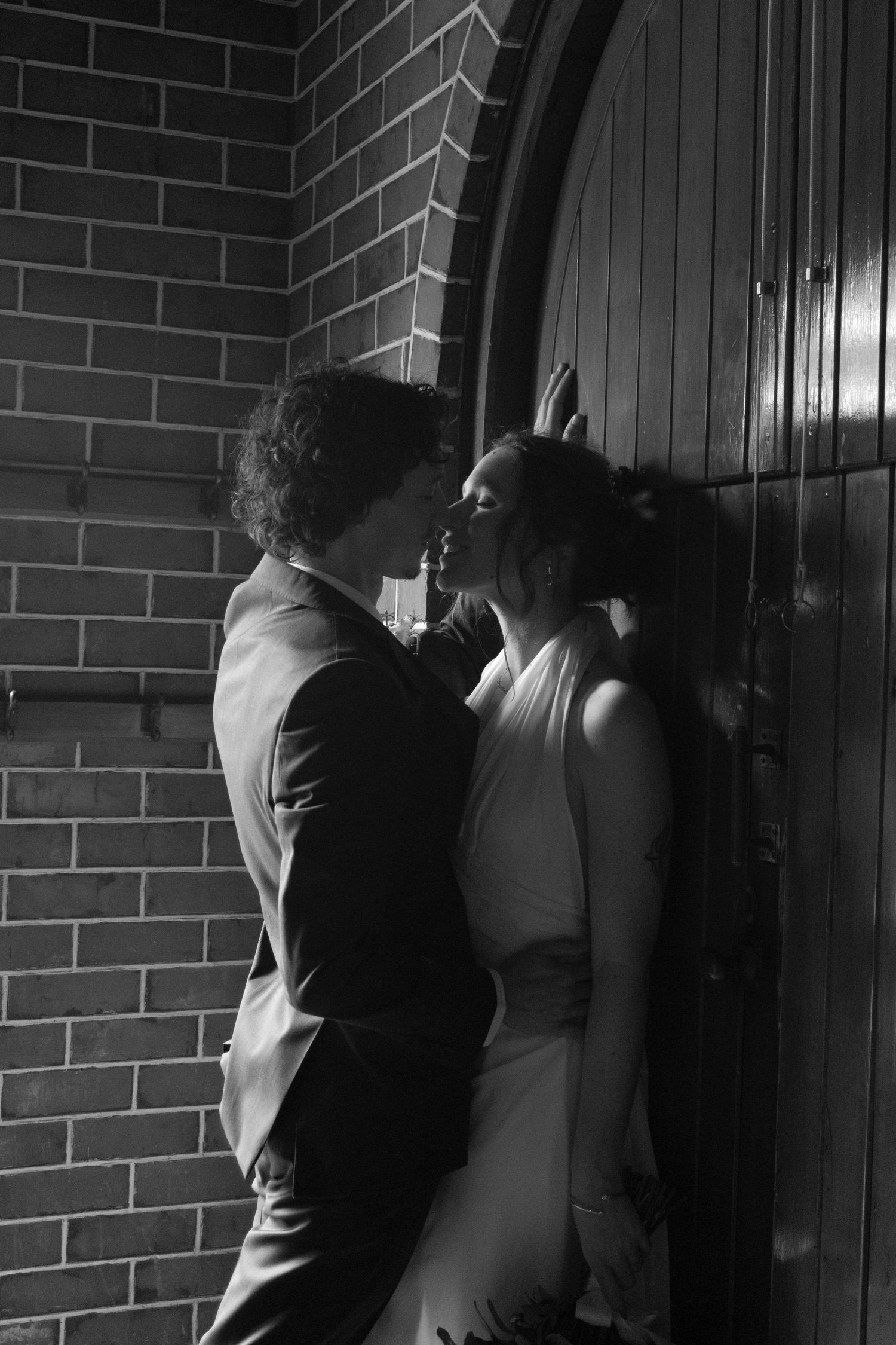 Black and white photo of a couple sharing a romantic kiss by a wooden door against a brick wall.