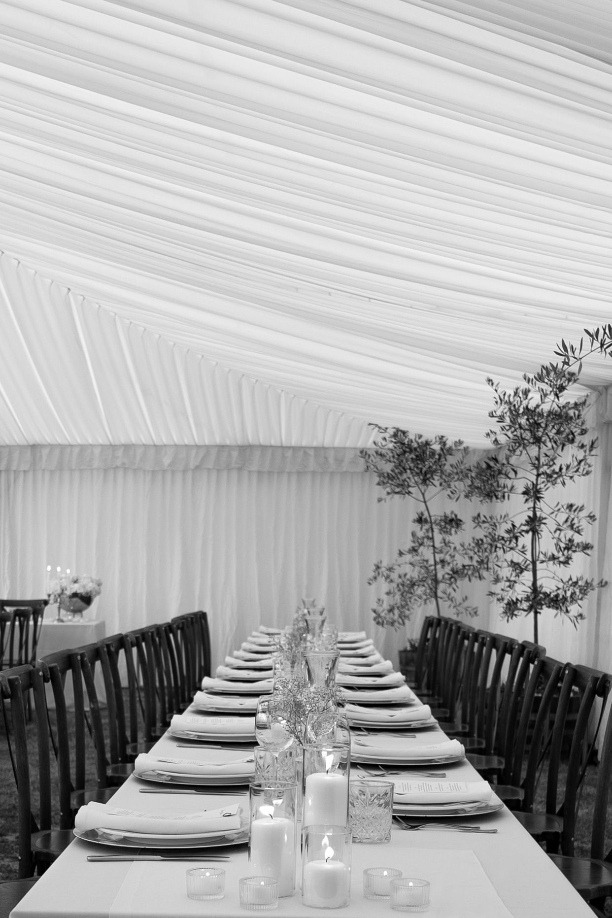 Elegant black and white photo of a long banquet table set with plates, candles, and glassware inside a draped marquee tent.