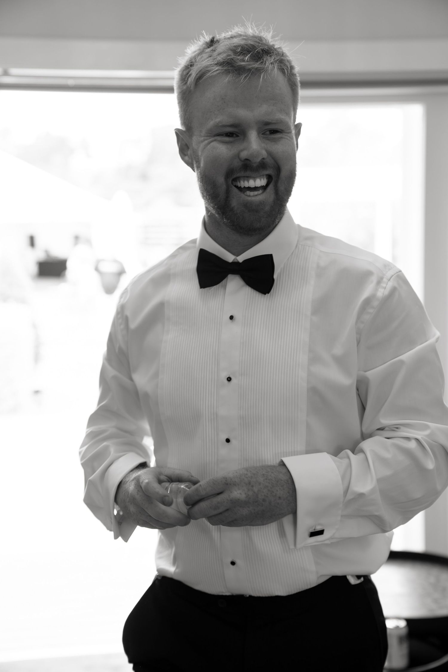 Smiling groom in white tuxedo shirt and black bow tie adjusting cufflinks before wedding ceremony.
