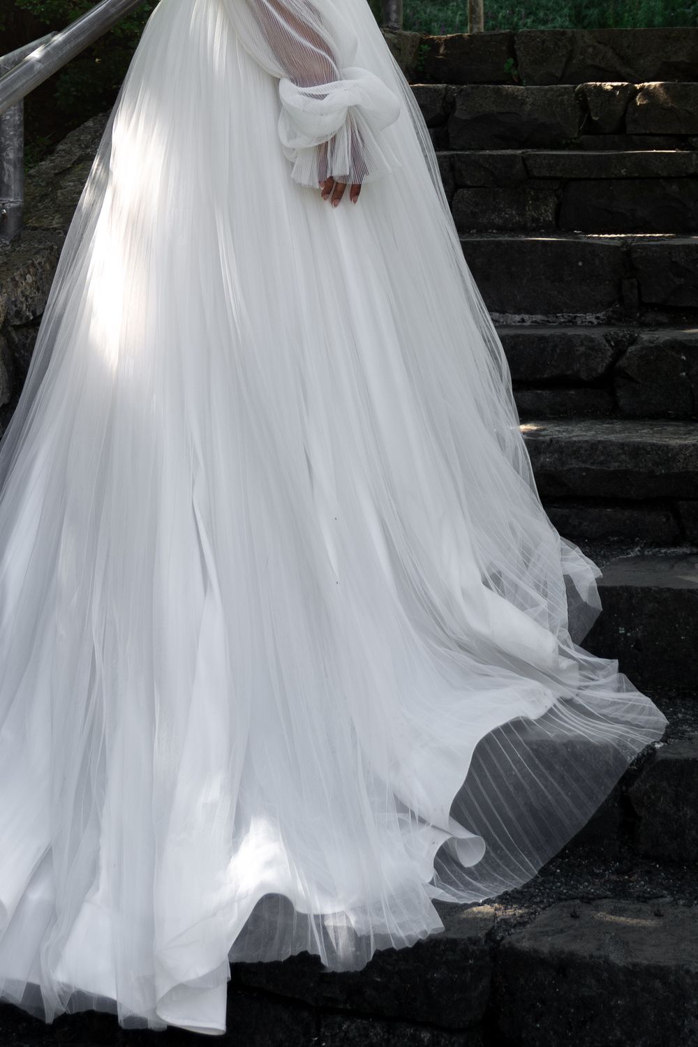 Bride in flowing white pleated wedding gown with long veil walking down stone steps outdoors.