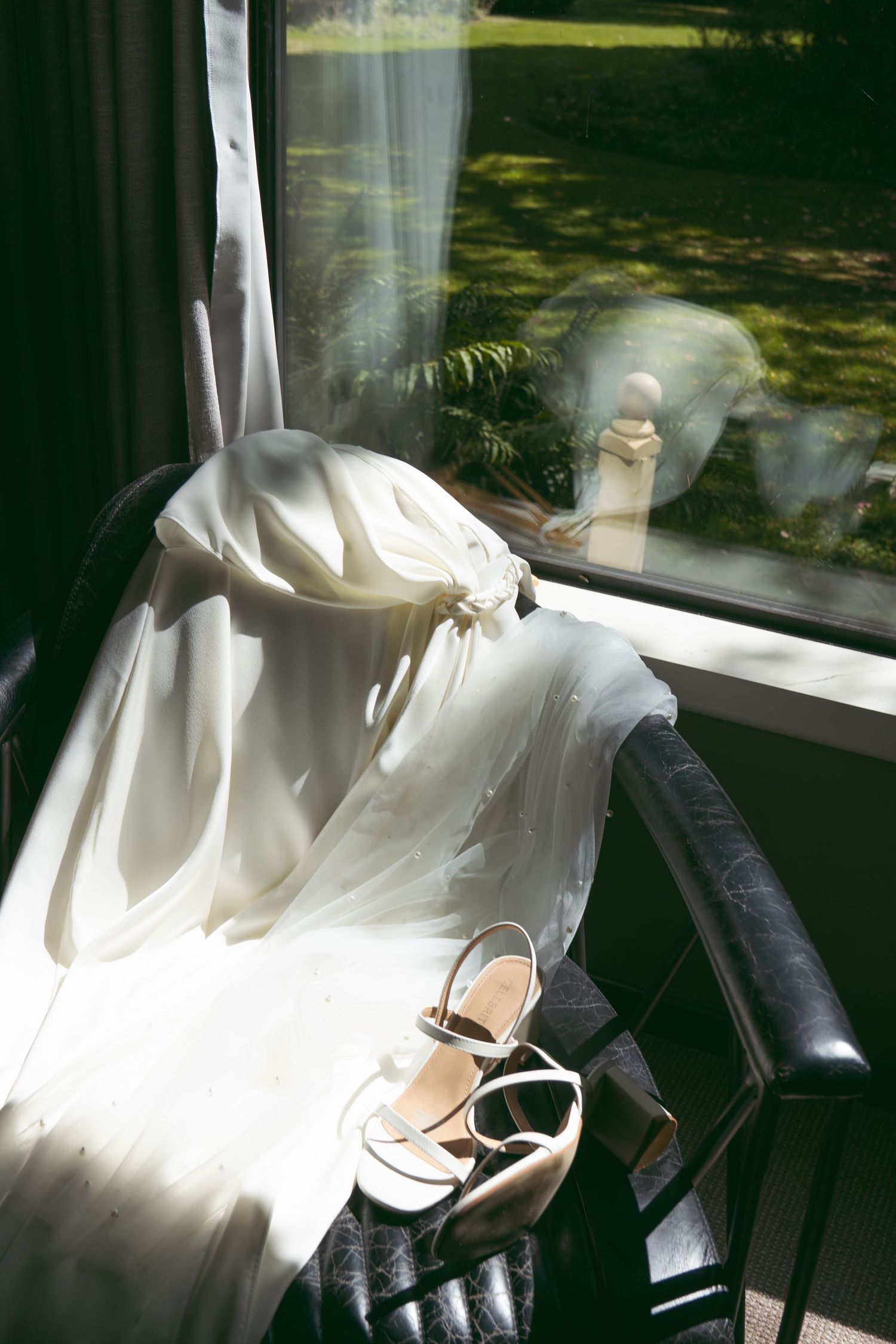 White bridal gown draped over a leather chair with nude heeled sandals beside a sunlit window.