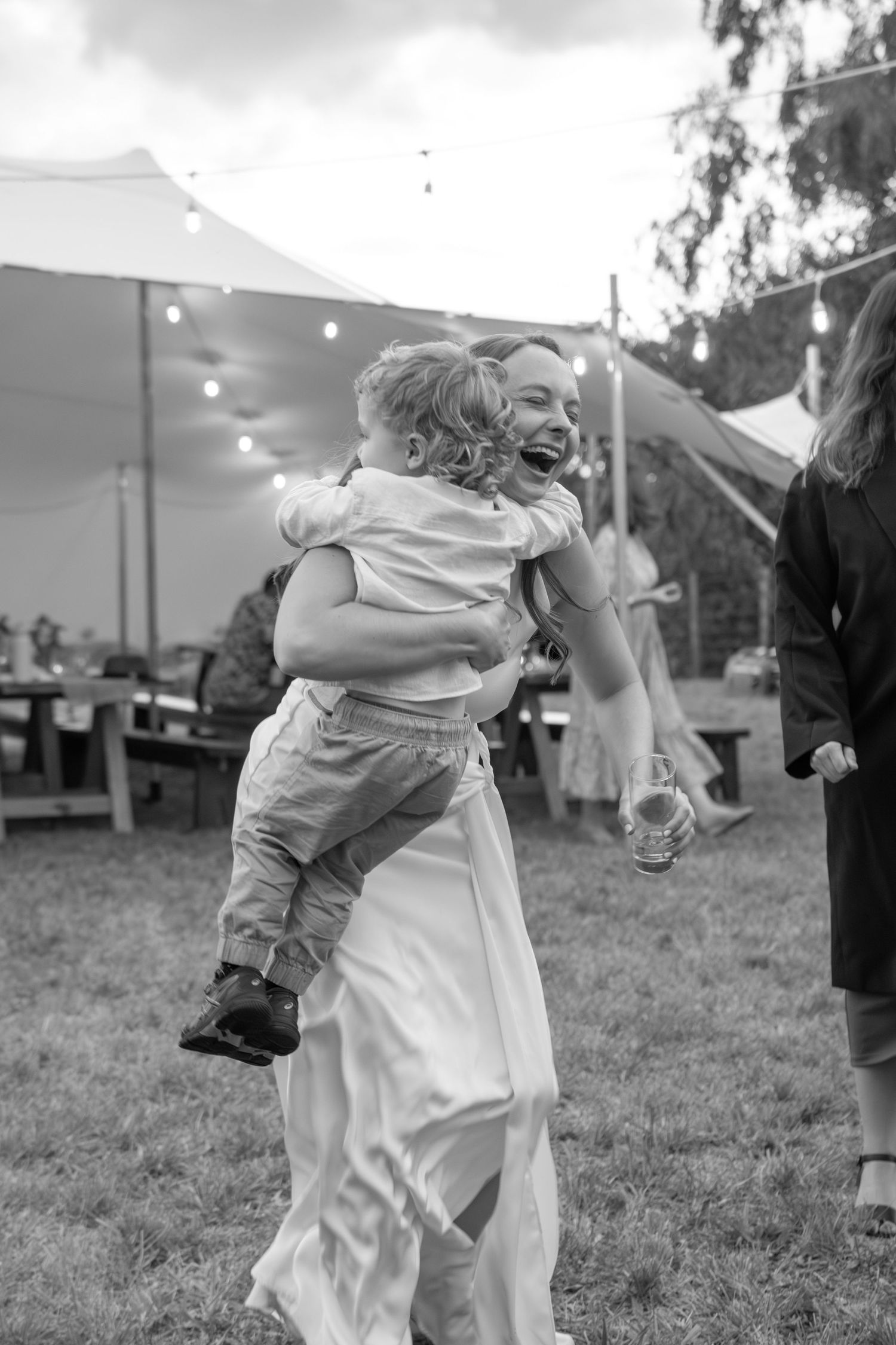 Black and white photo of a laughing bride holding a young child at an outdoor wedding reception with string lights.