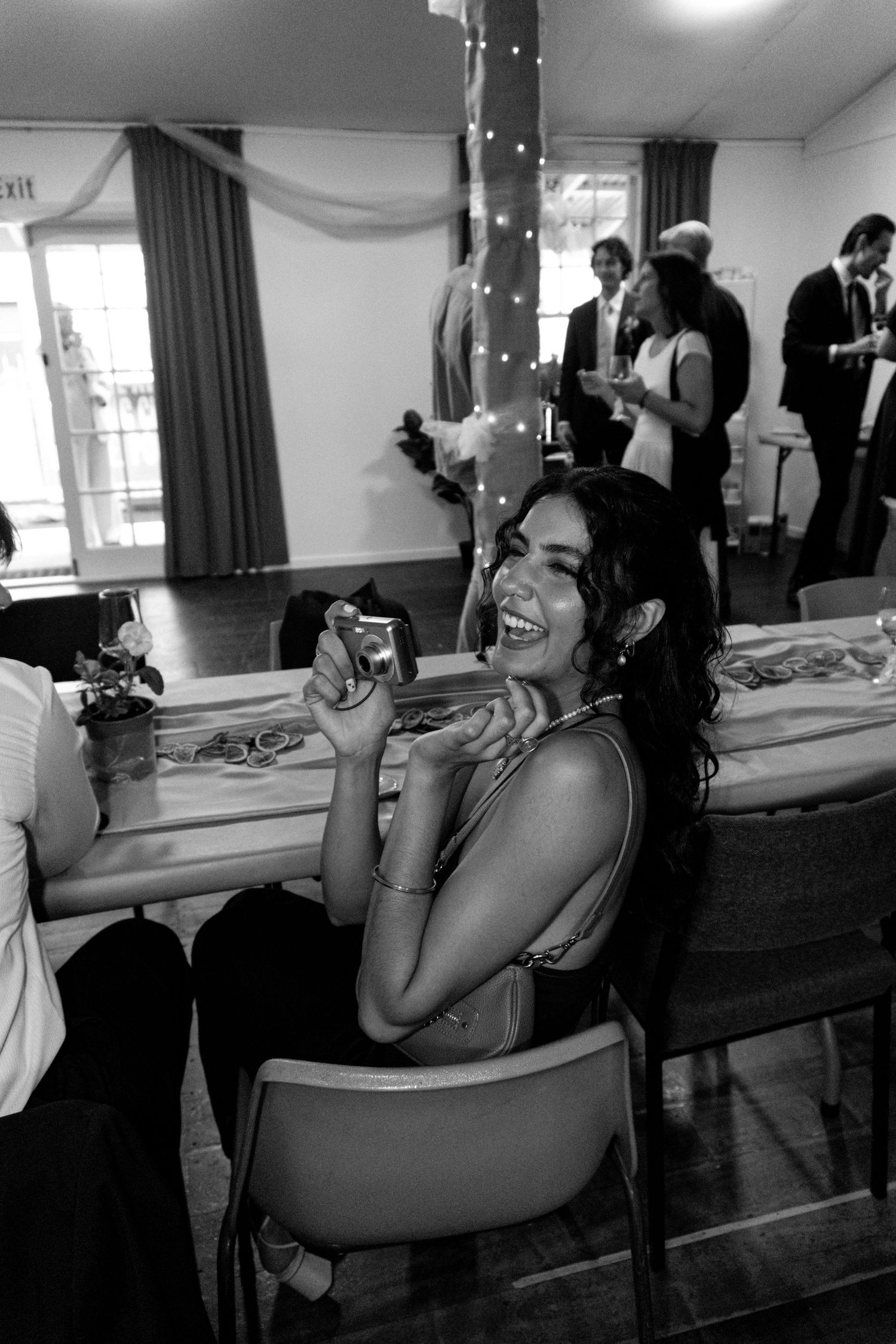 Black and white photo of a laughing woman holding a camera at a wedding reception, with guests socializing in the background.