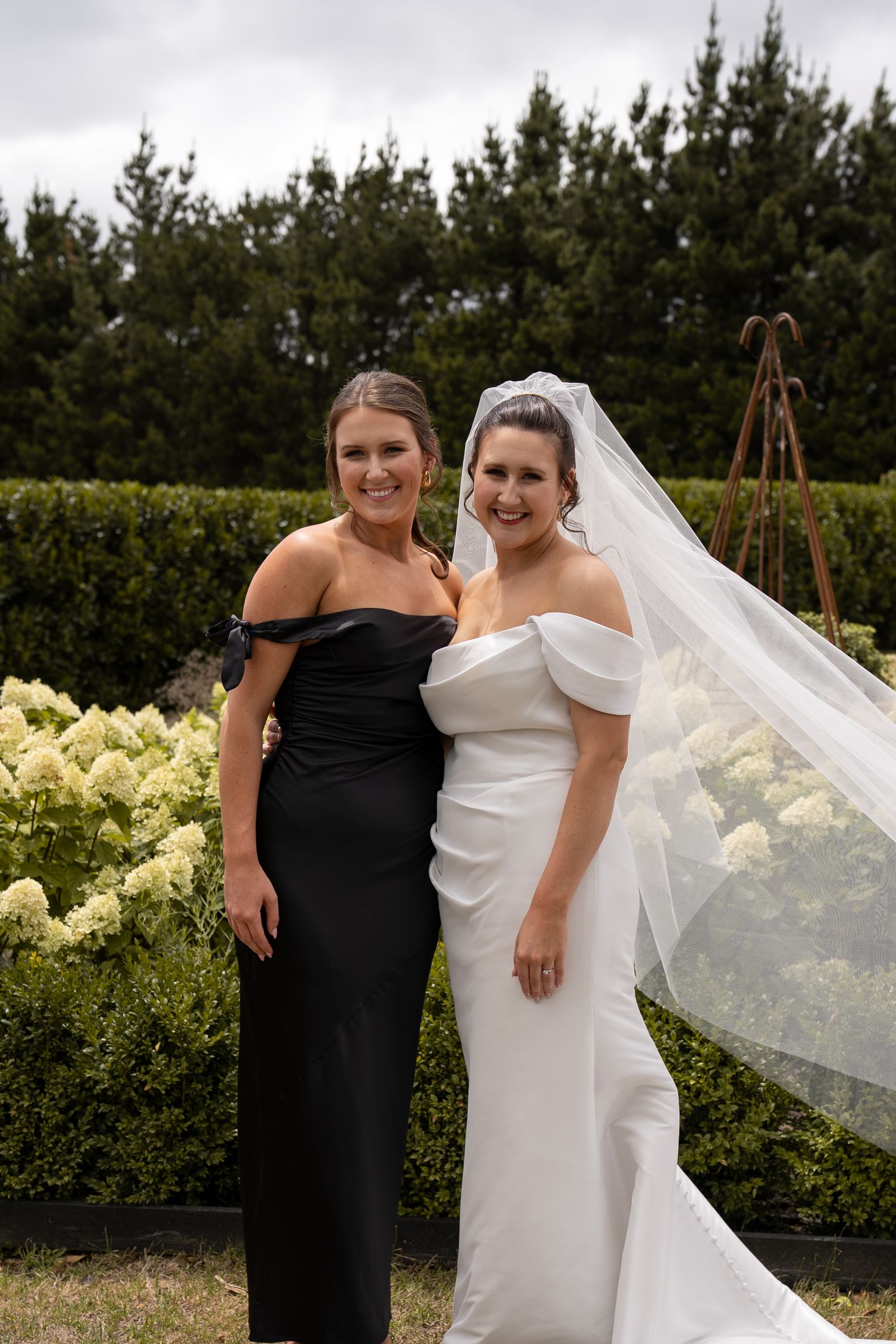 Bride in white off-shoulder gown with veil poses with bridesmaid in black dress in garden setting.