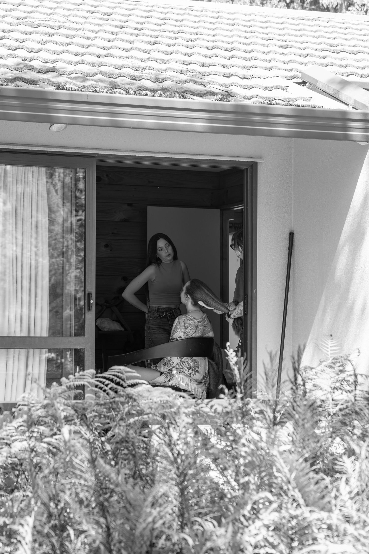 Black and white photo of two women standing in an open sliding glass door of a house surrounded by lush greenery.