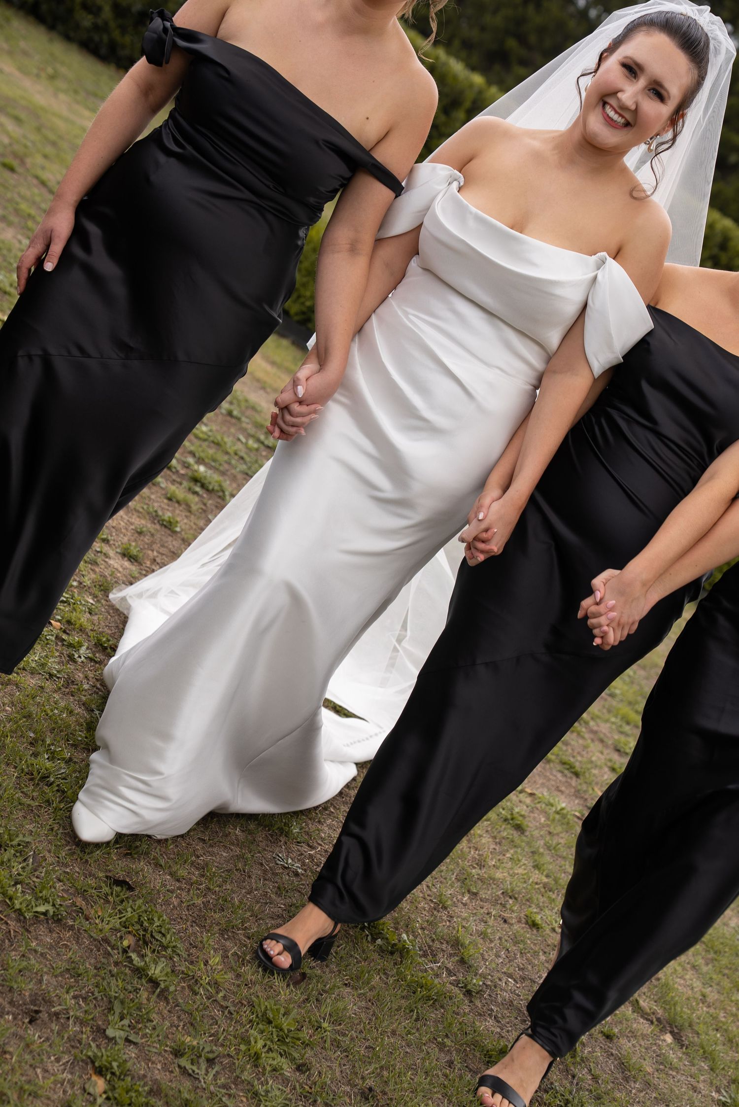 Bride in white off-shoulder gown holds hands with bridesmaids in black dresses outdoors.