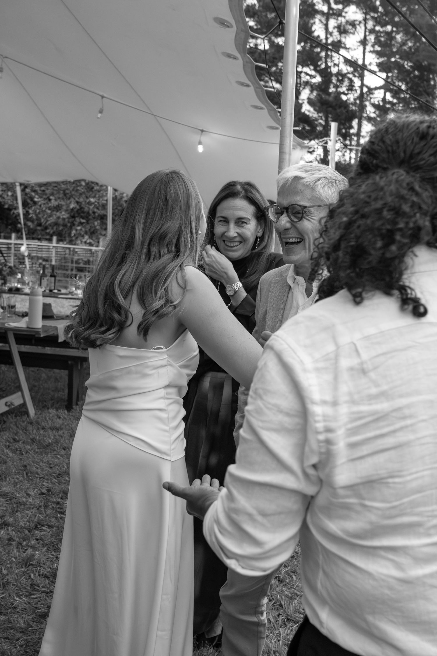 Black and white photo of a bride in white dress embracing guests under a tent at an outdoor wedding reception.