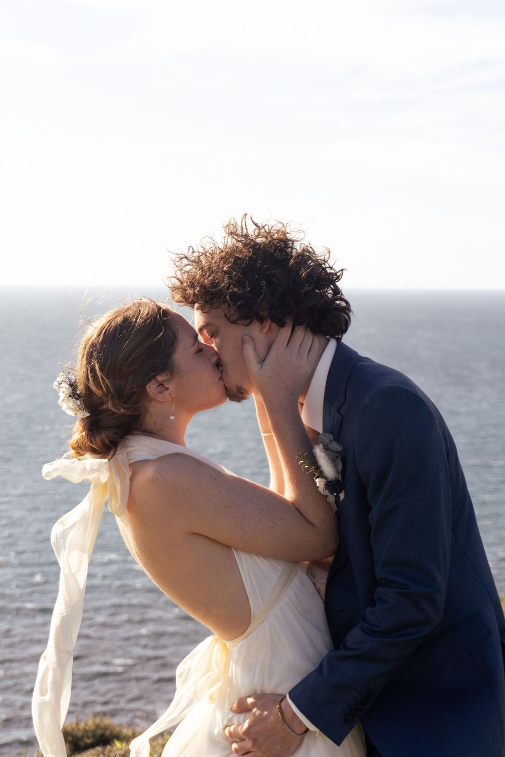 Bride and groom share a romantic kiss by the ocean on their wedding day, dressed in white gown and navy suit.