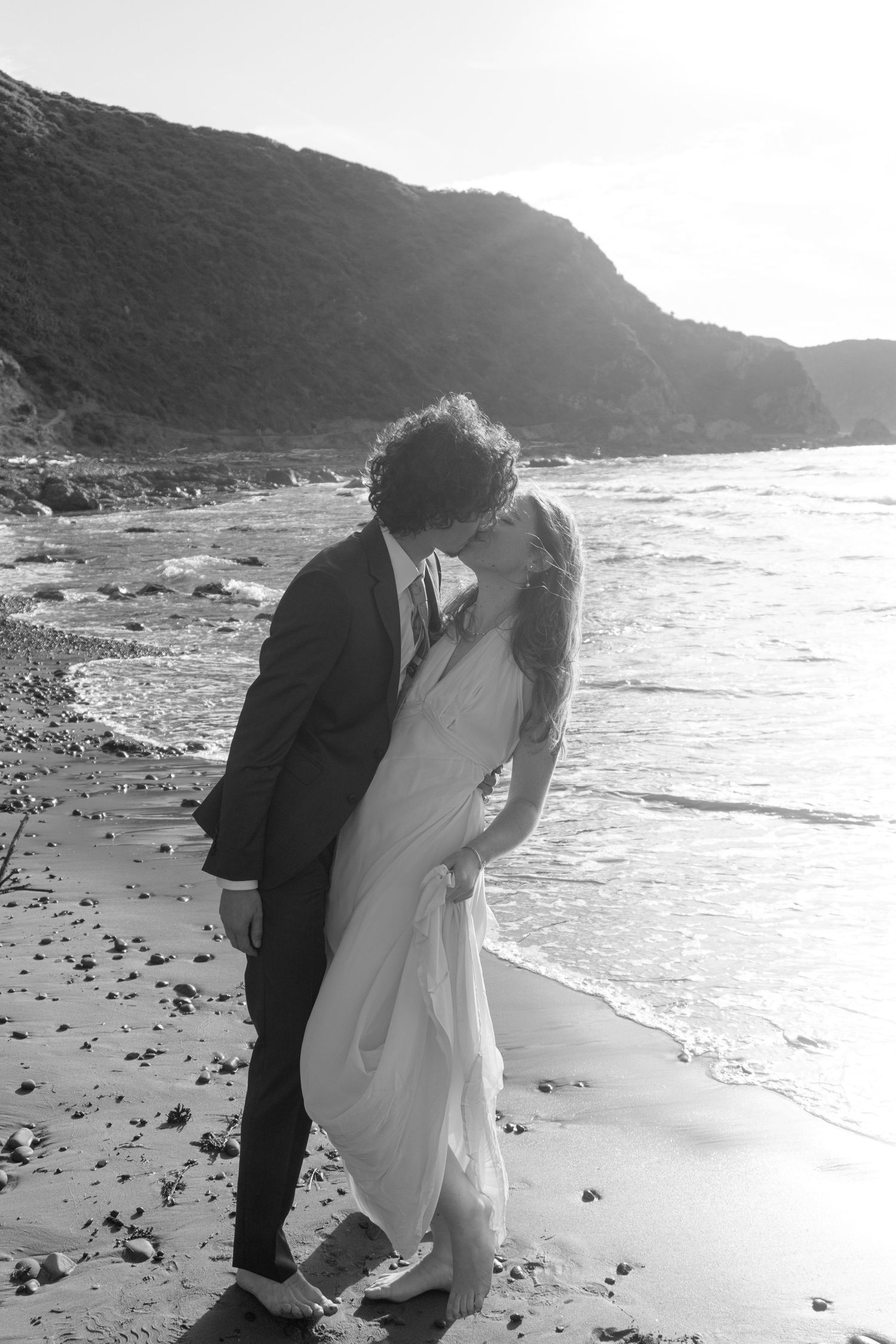 Black and white photo of a bride and groom kissing on a rocky beach with ocean waves and cliffs in the background.