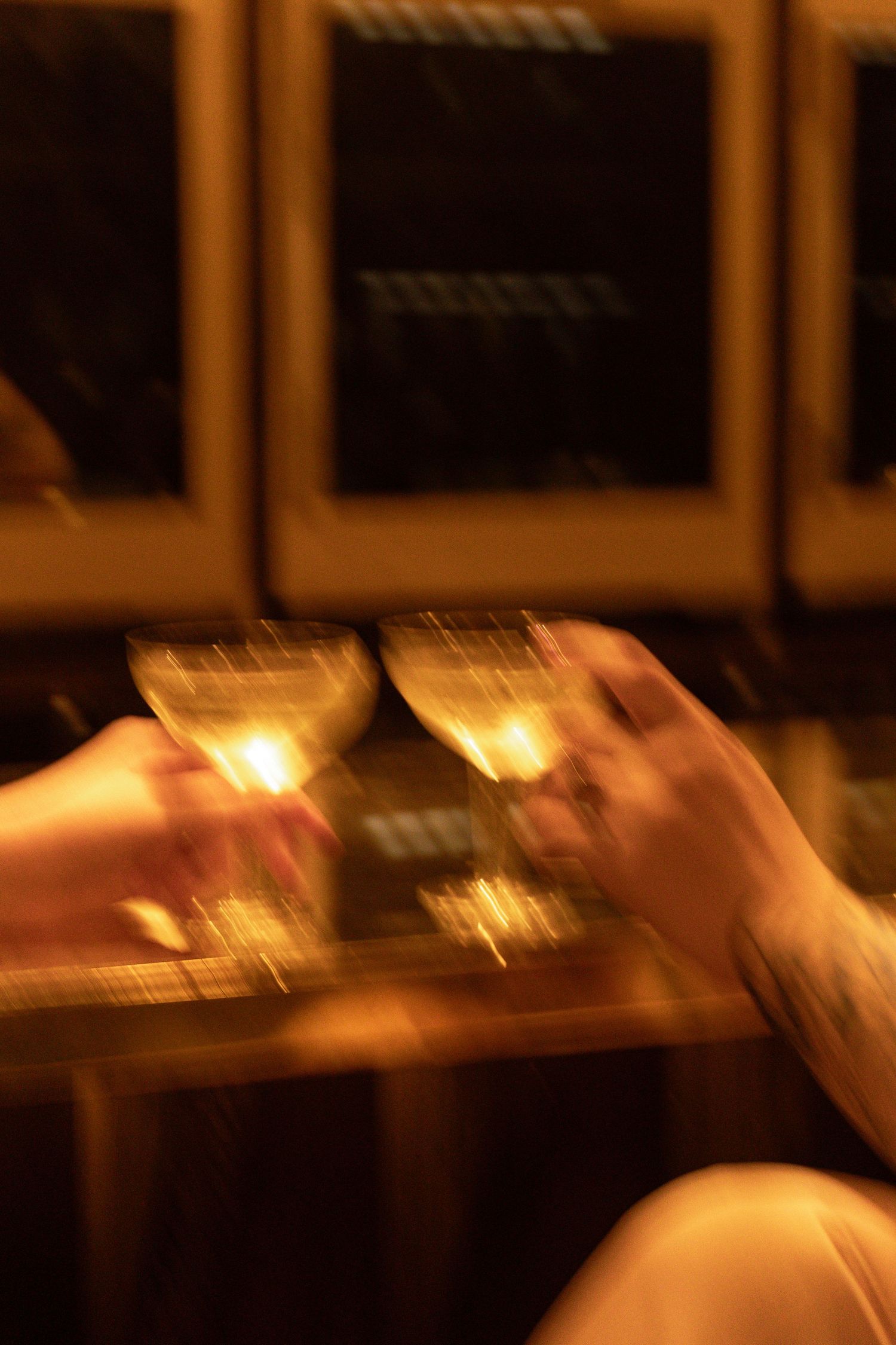 Two hands clinking wine glasses in a warm, candlelit bar setting for a celebratory toast.