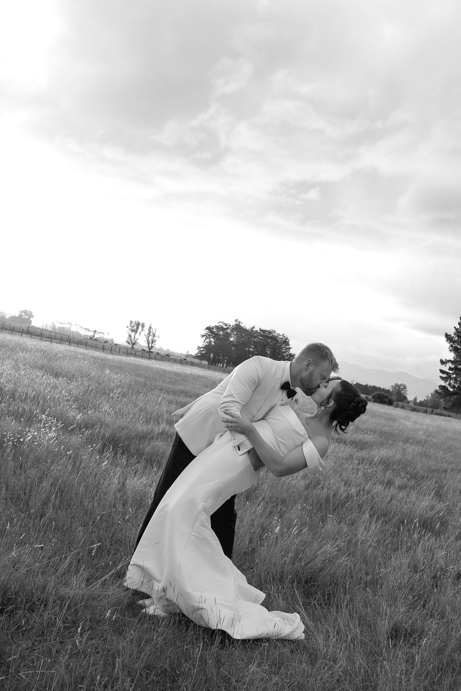 Black and white photo of a groom dipping his bride for a kiss in a grassy field on their wedding day.