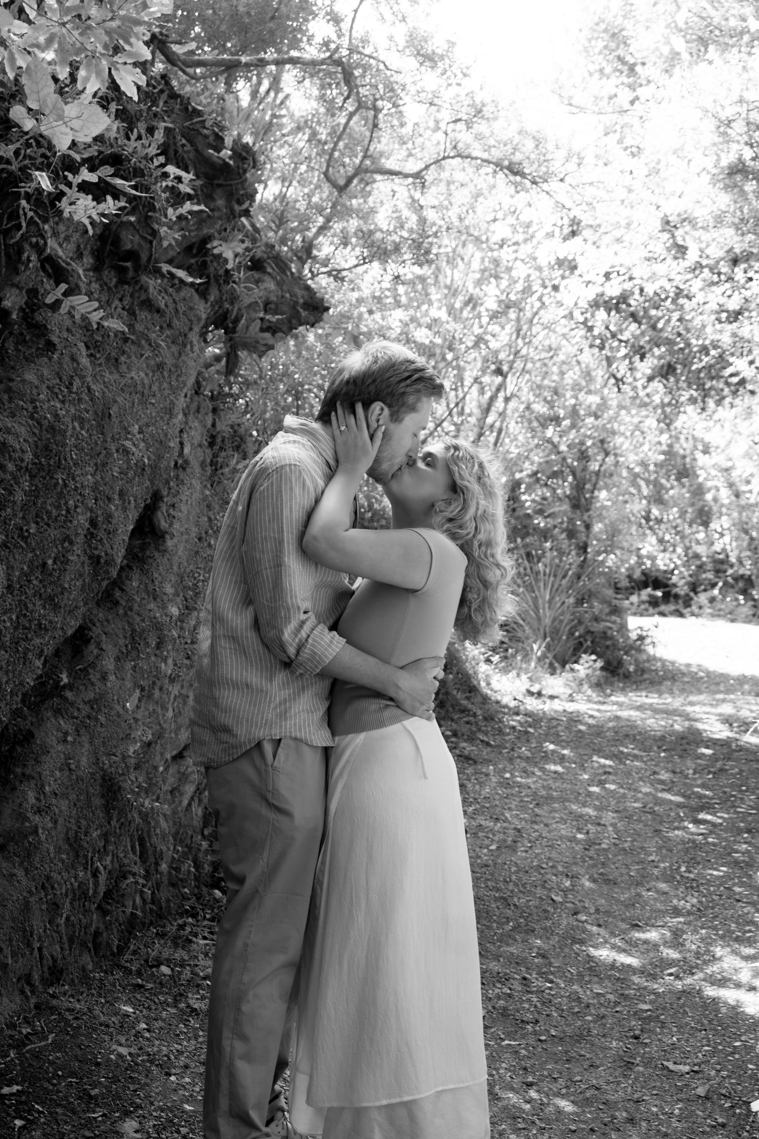 Black and white photo of a couple kissing tenderly in a lush forest setting, the woman in a flowing skirt embracing the man.