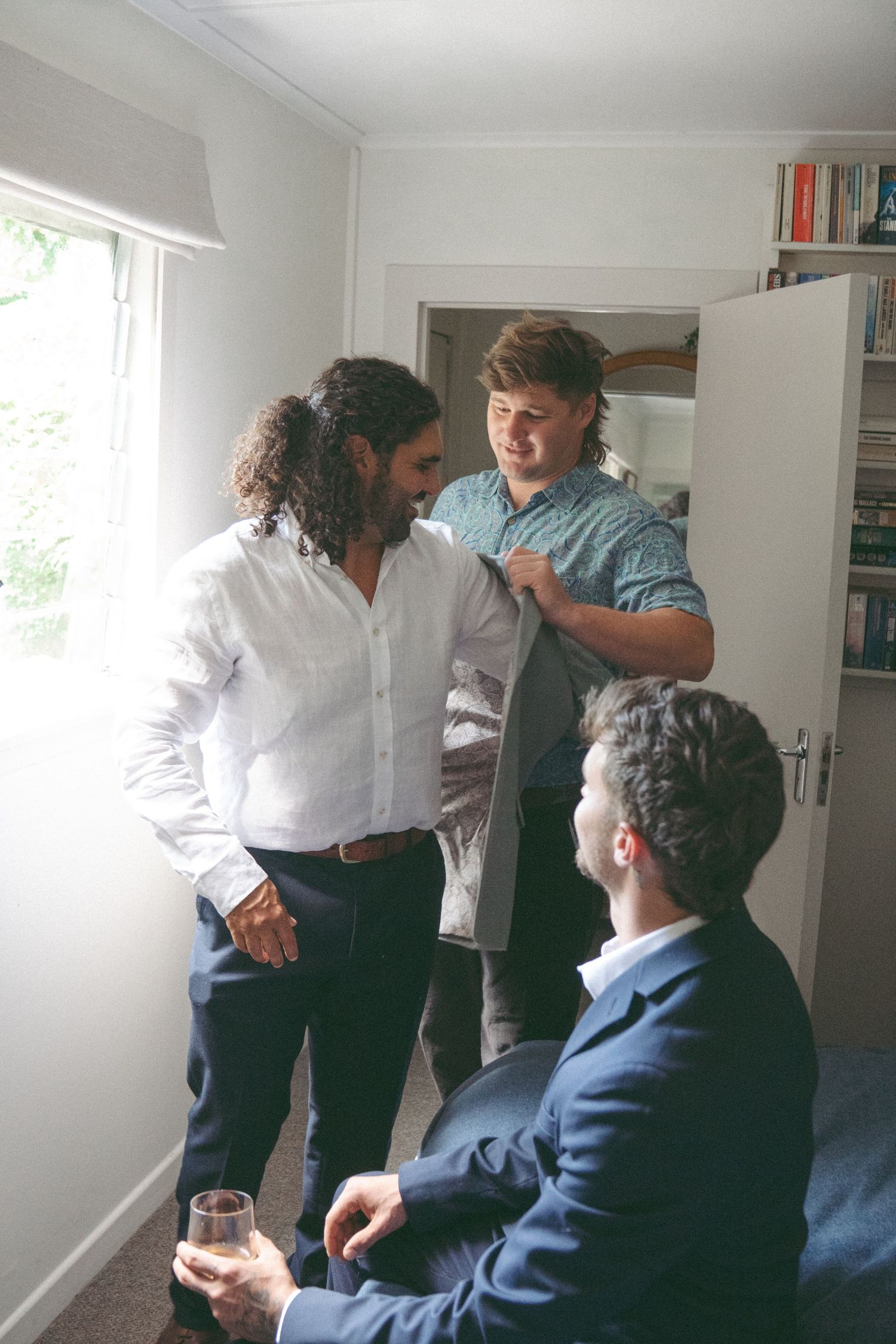 Three men in formal attire chat in a bright room, one holding a drink while another adjusts a jacket.