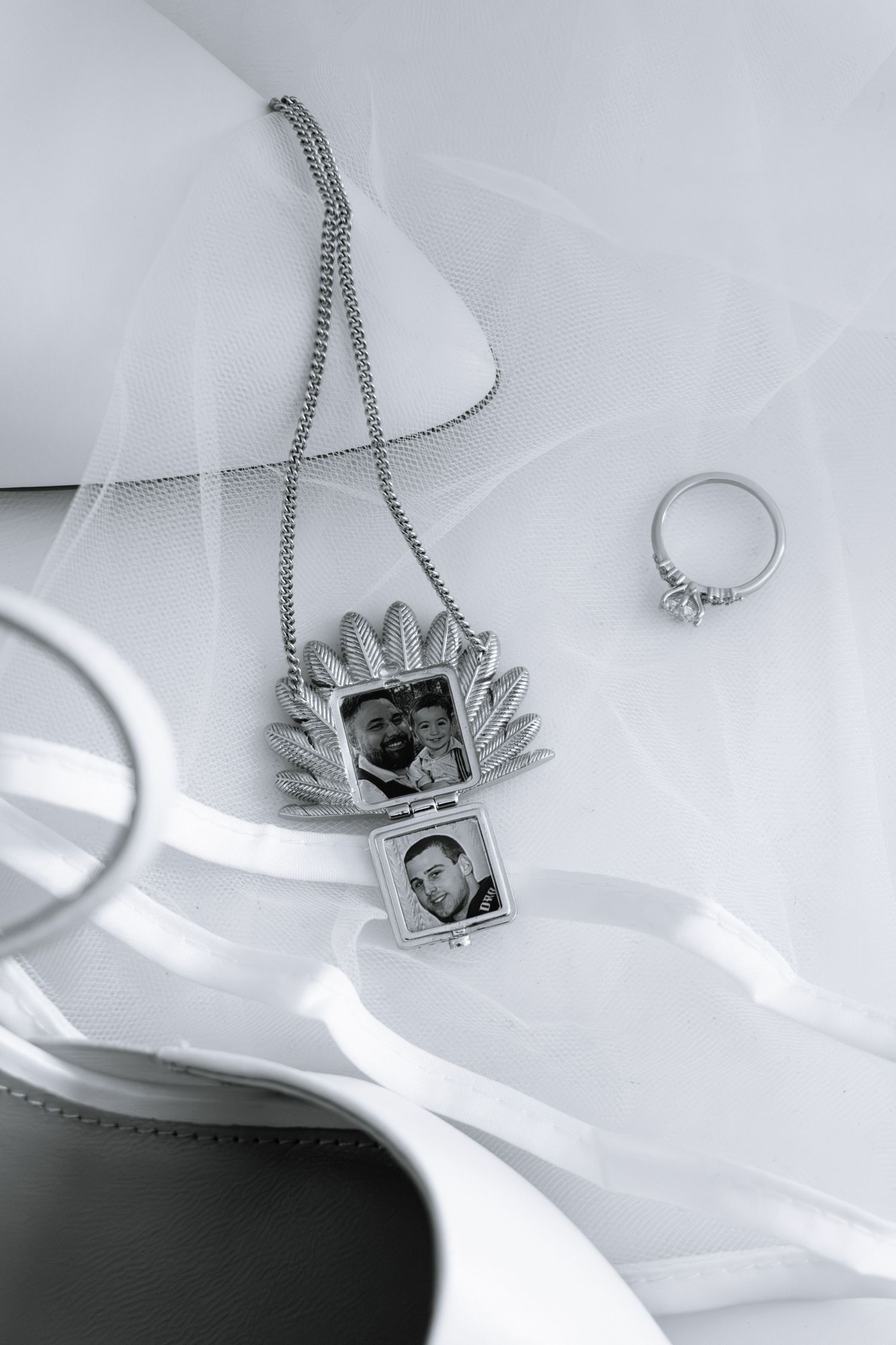 Silver locket necklace with family photos, diamond ring, and white ribbon on elegant bridal flat lay display.
