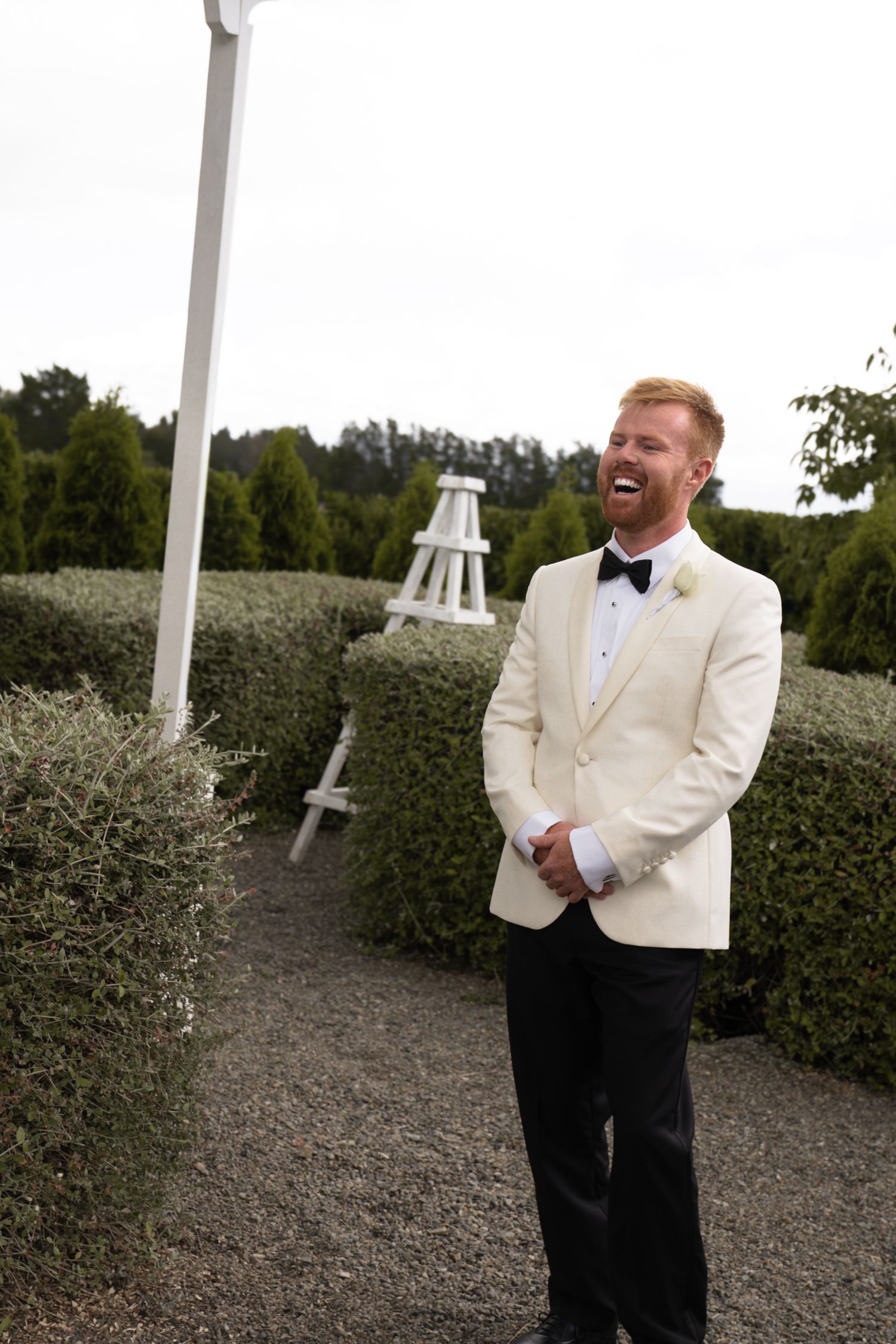 Laughing groom in white tuxedo jacket and black bow tie stands on gravel path surrounded by manicured hedges.