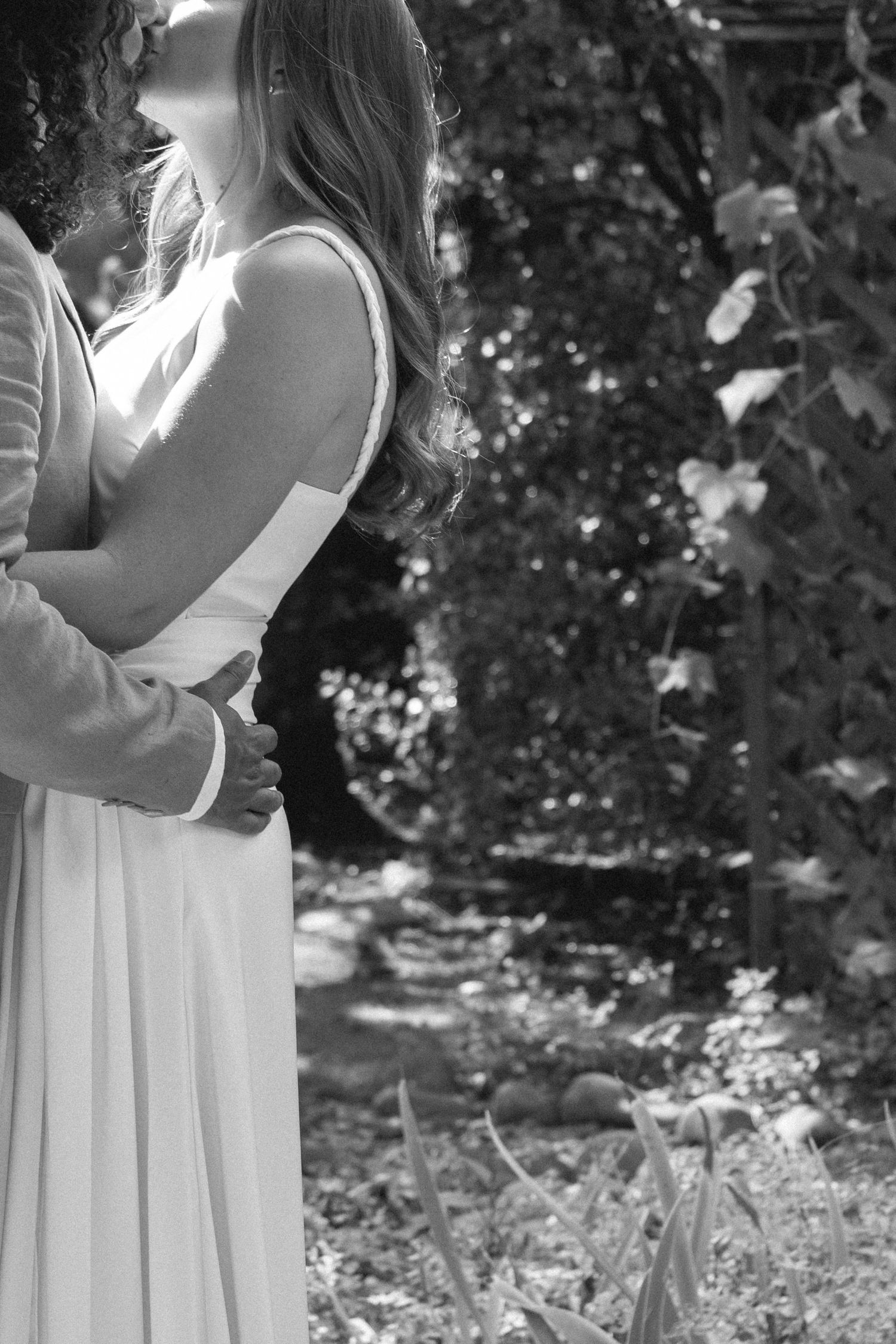 Black and white photo of a couple embracing outdoors, woman in white dress, surrounded by lush garden foliage.
