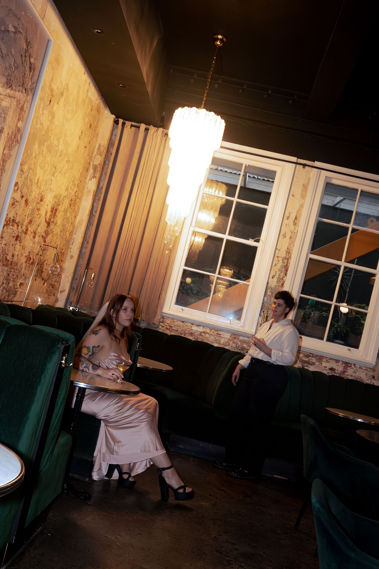 Two elegantly dressed people conversing in a dimly lit upscale lounge with green velvet seating.